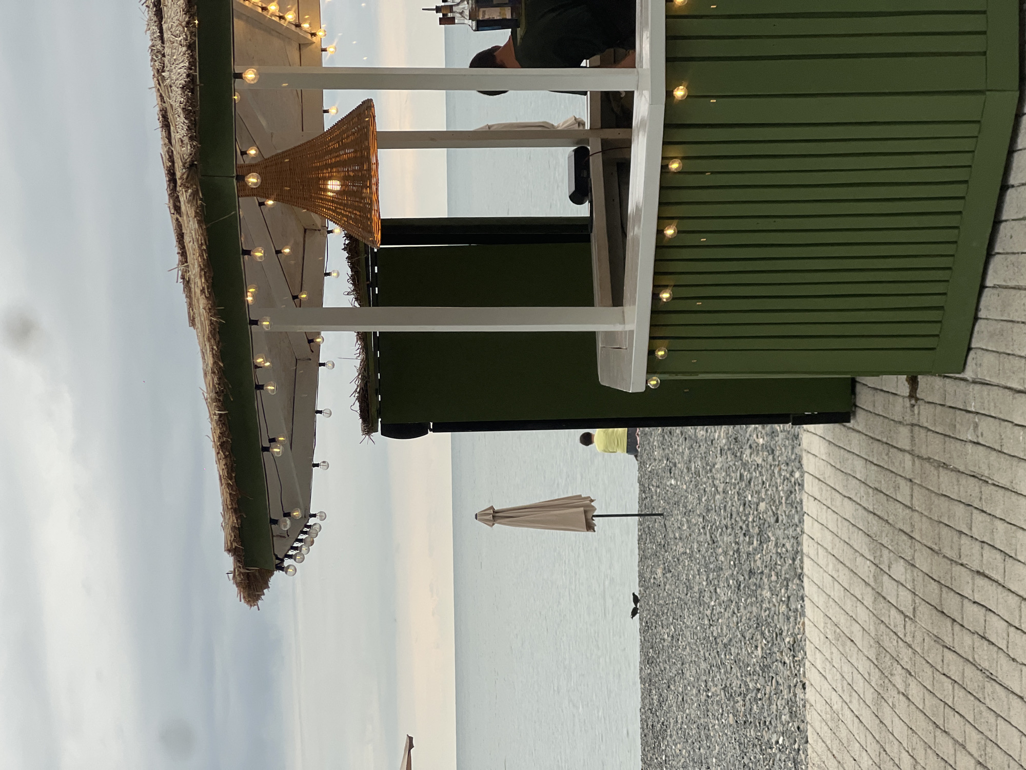 A low-angle, eye-level outdoor shot depicts a beachside bar in Sokhumi, Abkhazia, on an overcast day. The bar, painted olive green, features a thatched roof adorned with a string of small, warm-toned lights. A woven pendant light hangs in the center of the bar area. The bar counter is white, and more string lights are visible below the counter. A person in a dark shirt is behind the counter, appearing to work. In the background, the calm, grey sea stretches to the horizon under a similarly grey sky. The beach is covered in dark grey pebbles. A closed, beige beach umbrella stands upright on the pebbles. Two dark birds are on the beach near the umbrella, and a person in a bright yellow shirt sits further down the beach, facing the sea. A paved pathway, made of interlocking rectangular stones, occupies the foreground.