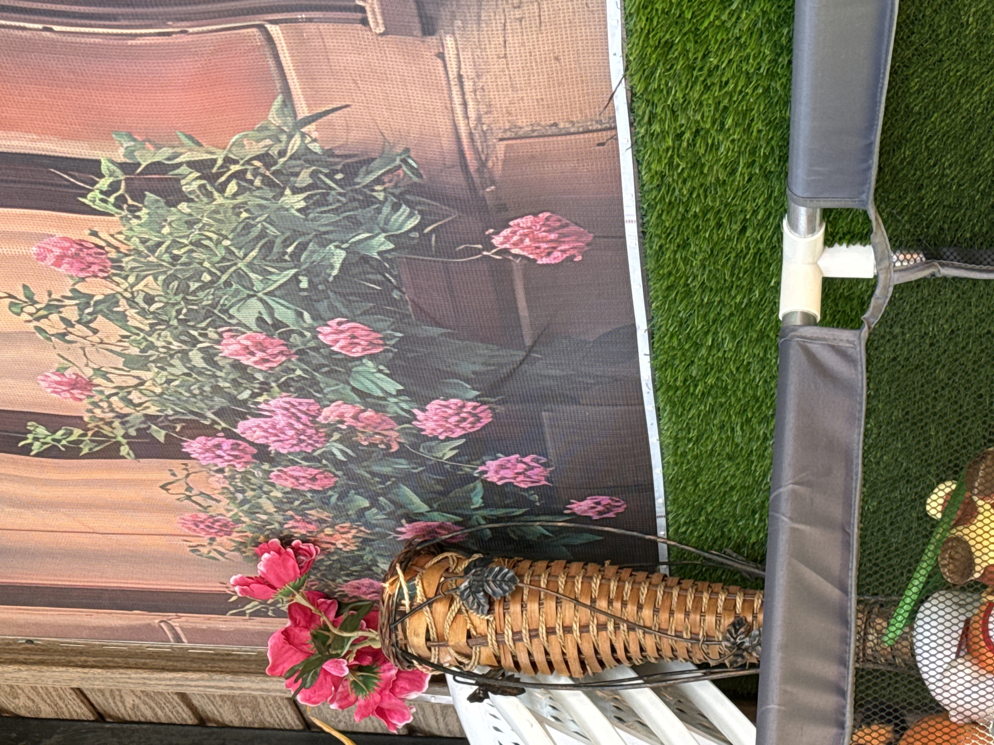 The image shows a close-up of a floral arrangement against a backdrop that appears to be a printed image or canvas depicting a building facade with flowers. The dominant colors are pink, green, brown, and gray.
The floral arrangement consists of pink flowers and green foliage. It is partially situated in front of a woven basket or container.
To the right, a section of what looks like a padded or fabric-covered frame is visible, positioned against an area of artificial green turf.
No people, specific actions, or interactions are evident. The location is not discernible.
