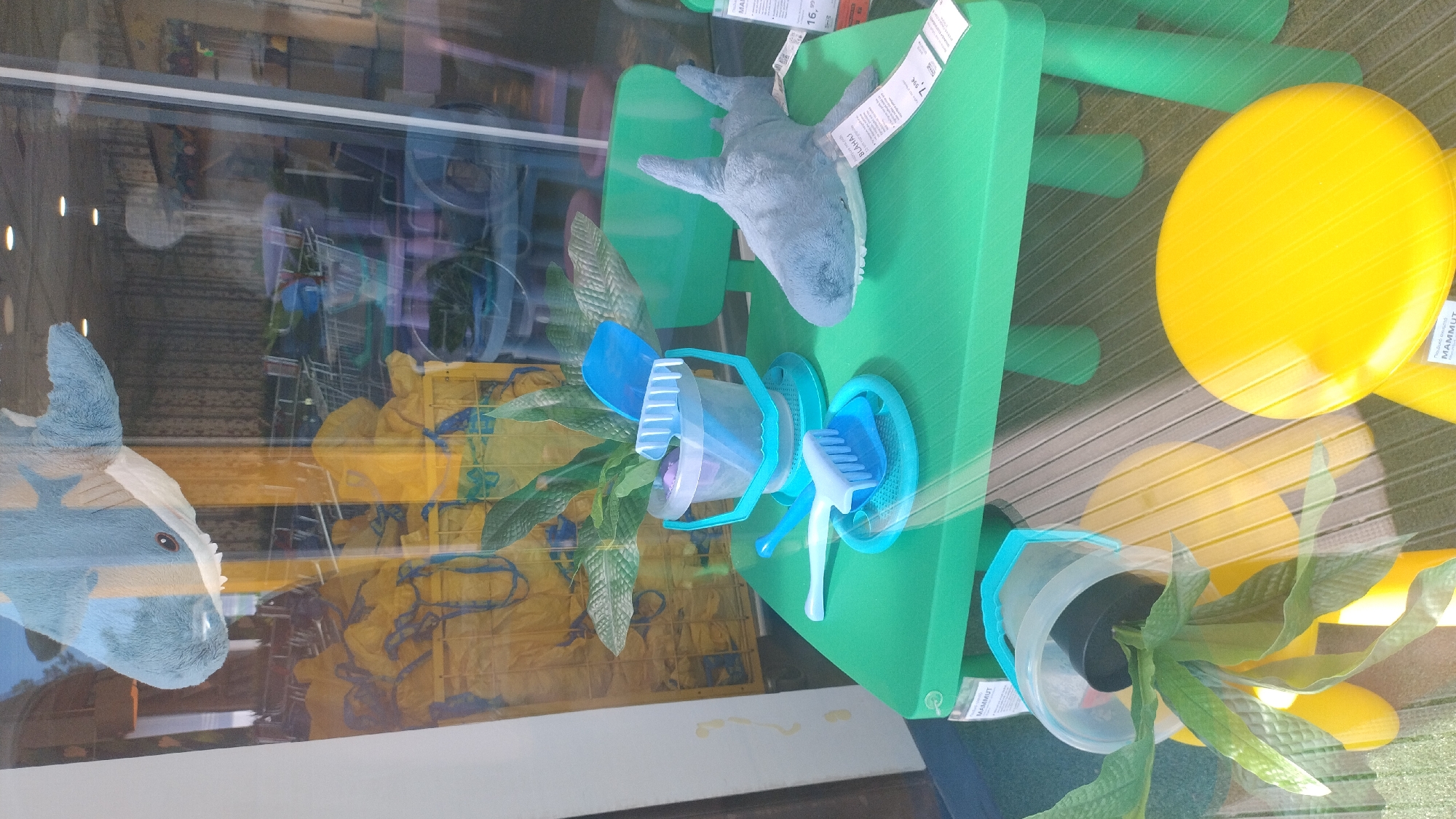 This media file shows a retail display, likely within a store, reflecting its surroundings. The primary subjects are several IKEA MAMMUT products: a green stool, a yellow stool, and a small blue and clear plastic container with a brush. A plush gray shark toy is placed on top of the green stool, and a plush blue toy, possibly a unicorn or horse head, is partially visible on the left. A green plant in a clear plastic pot sits on the floor in front of the yellow stool. The reflection in the glass reveals more of the store's interior, including shopping carts, shelving, and yellow plastic crates filled with merchandise.
The scene appears to be set indoors during daylight hours, with natural light creating reflections and highlights on the glass and products. The overall impression is of a clean and organized retail environment showcasing children's furniture and toys. The reflection suggests the display is near a window or glass storefront. The only visible text is on the product tags, which appear to be IKEA labels. The "MAMMUT" branding is legible on at least two of these tags. The context of Pylaia Municipal Unit, Greece, places this scene in a specific geographical location. No specific activity or event is taking place, other than the display of products.
