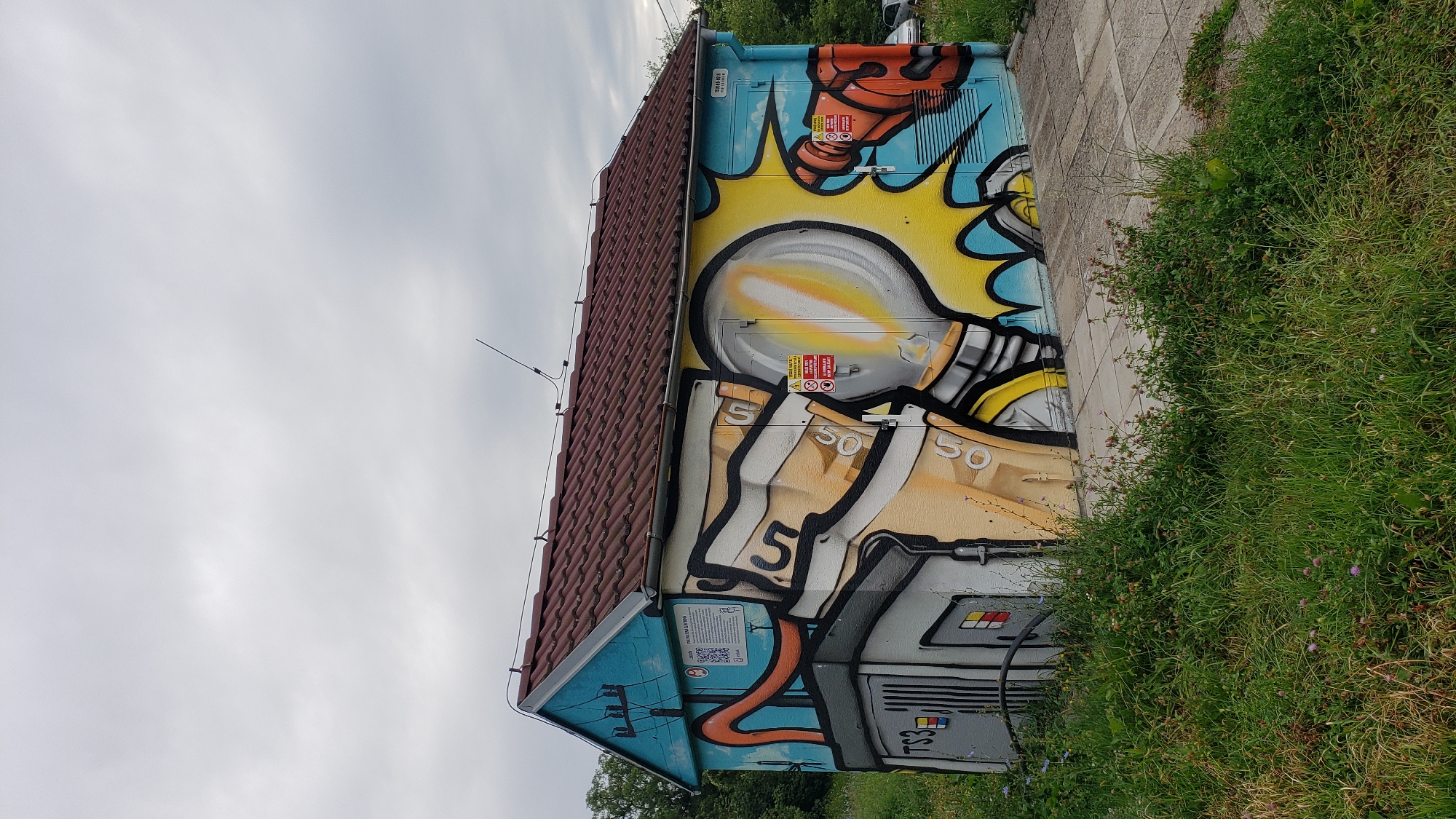 The image shows a small, rectangular building with a brown tiled roof, covered in vibrant graffiti art. The graffiti depicts a large, stylized lightbulb with a yellow glow, a hand holding a plug, and what appear to be electrical components. The building itself is painted light blue on the left side, with a portion of the graffiti extending onto this side as well. The right side of the building is predominantly yellow and blue, with the main lightbulb artwork. There are several warning signs with red and white symbols visible on the building's doors. The building is situated on a paved pathway surrounded by overgrown green grass and some scattered purple wildflowers. In the background, there are trees and a cloudy sky. A car is partially visible in the far right background. The building is likely an electrical substation or utility box. The graffiti art suggests a creative and artistic beautification of an otherwise functional structure. This scene is located in the city of Prešov, Slovakia.