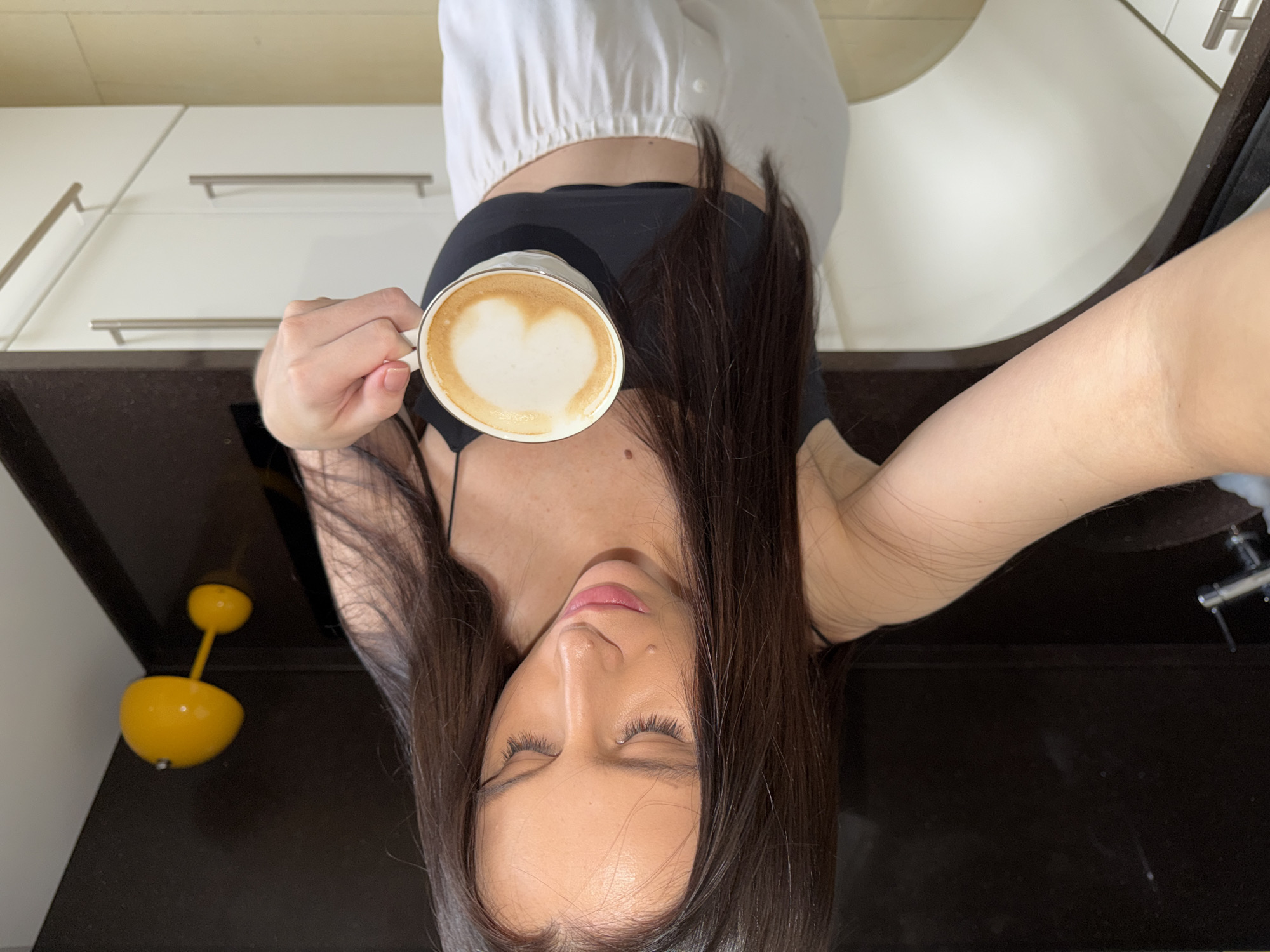 A woman in a black top and white pants holds a cup of coffee with a heart-shaped latte art in a kitchen. The background includes kitchen cabinets, a black countertop with a yellow fruit bowl, and a sink with a faucet. The location is Khimki, Russia.