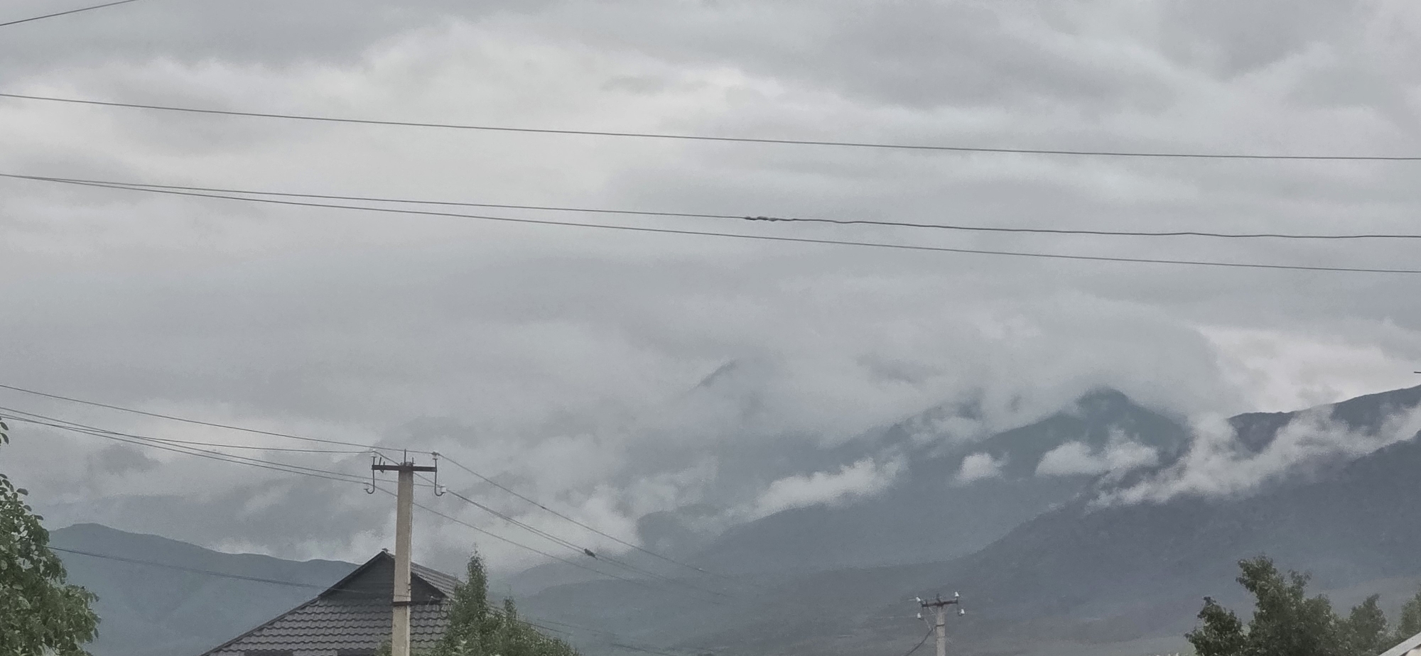 The image depicts a cloudy, overcast day in Chong-Sary-Oy, Kyrgyzstan, with a focus on the mountainous landscape. Several power lines traverse the frame horizontally, supported by utility poles. In the foreground, the dark, tiled roof of a building is partially visible on the left, along with some green foliage. The middle ground is dominated by layered, mist-shrouded mountains. The sky is filled with thick, grey clouds, obscuring any direct sunlight and contributing to a subdued atmosphere. The overall scene suggests a cool, possibly damp, day in a rural or semi-rural mountainous region.