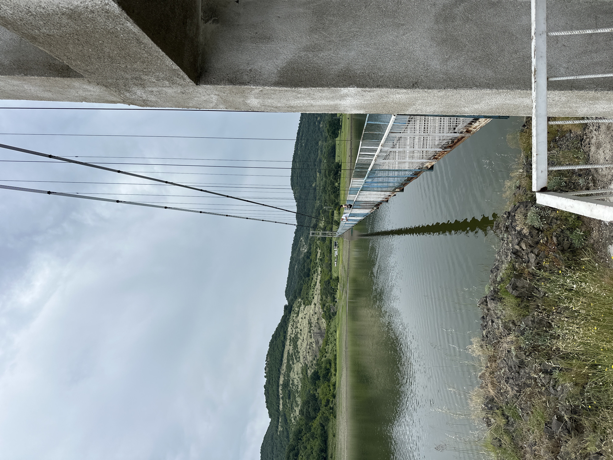 This image depicts the Lisitsite suspension bridge in Lisitsite, Bulgaria, extending over a body of water, likely a reservoir, under an overcast sky. A prominent, unpainted concrete abutment occupies the right side of the frame, from which multiple metal suspension cables descend towards the bridge deck. The bridge deck consists of wooden planks flanked by weathered metal railings painted in blue and white, showing visible rust. Two individuals are observed on the bridge in the mid-ground: one closer, facing left, and another further along the span. The water's surface is rippled and reflects the bridge components. The left foreground reveals a rocky embankment with sparse dry grass and green foliage. Beyond the water, undulating hills covered in dense green vegetation and lighter, exposed patches form the background. Distant structures, possibly vehicles or small buildings, are faintly visible on the far shore. A section of white painted metal railing is present in the lower right, connected to the concrete abutment.