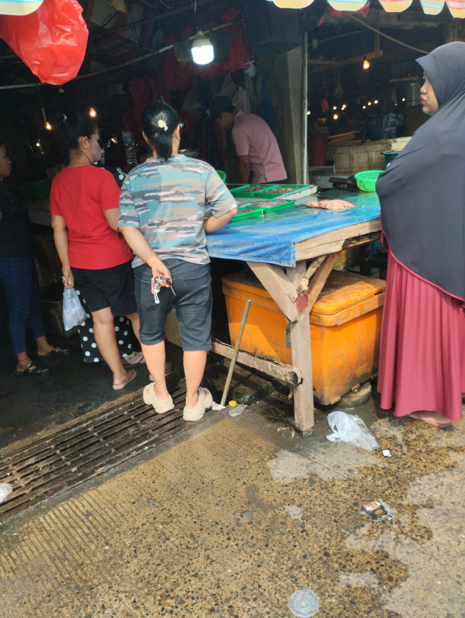 An outdoor market scene in Bekasi, Indonesia, shows several people interacting near a stall. A woman in a red shirt and black shorts stands holding a plastic bag. Beside her, a person in a camouflage shirt and denim shorts is reaching into their back pocket for keys. To the right, a woman wearing a black hijab and a long red skirt observes the scene. In the background, a person in a pink shirt is visible behind a table laden with fresh fish. The ground is wet and grimy, with a metal grate to the left and scattered debris, including plastic bags and what appears to be packaging, on the concrete floor. An orange cooler is visible under the table. A red plastic bag hangs from the ceiling in the upper left of the frame, and a bright light illuminates the area.