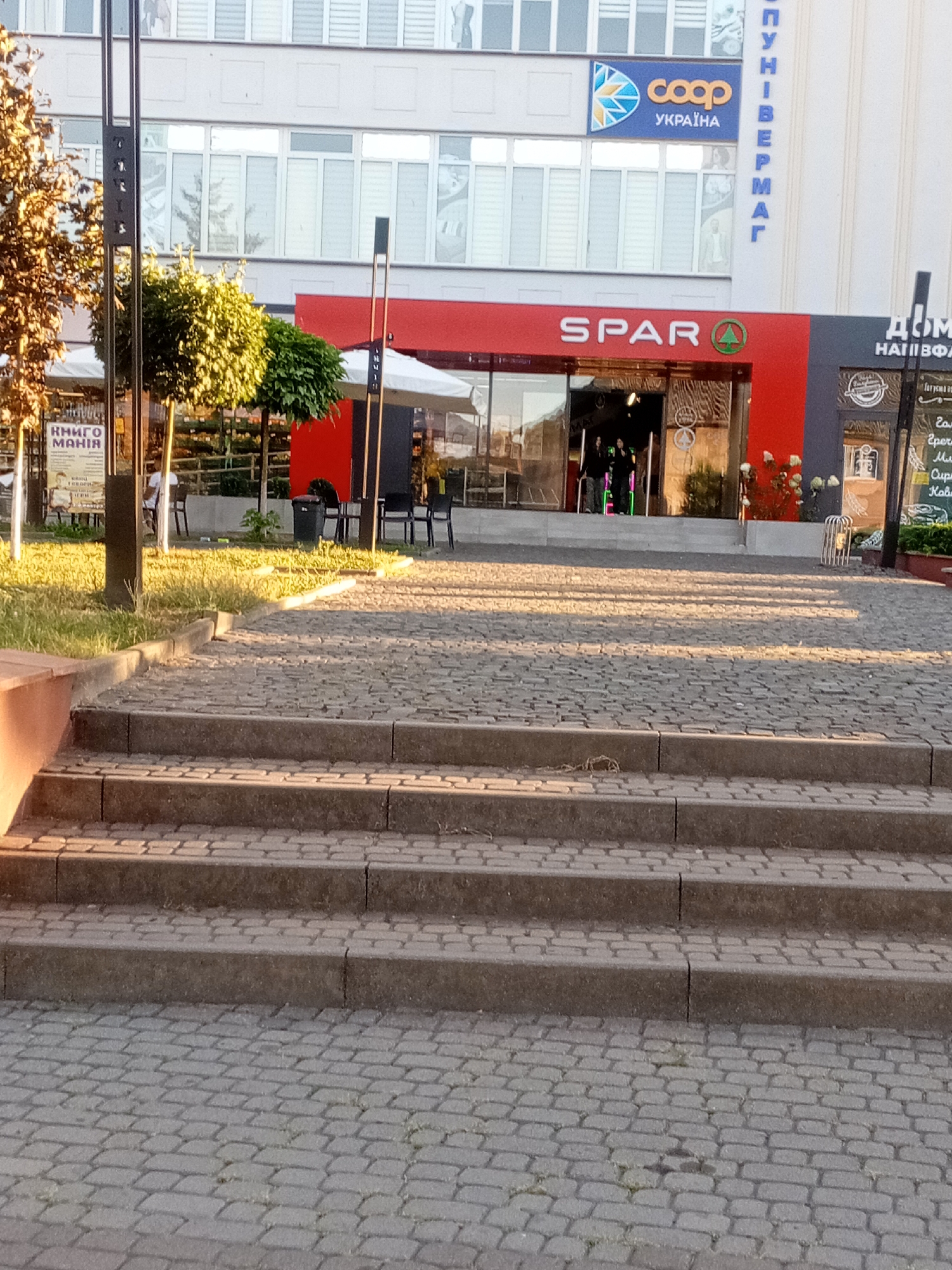 The image depicts the entrance of a SPAR supermarket in Tiachiv, Ukraine. The entrance is marked by a red awning and a glass facade. The SPAR logo is prominently displayed. To the left of the SPAR entrance, there is a sign for "КНИГО МАНІЯ" (Book Mania). Further to the right, a sign with the text "ДІМ" (House) is visible. The scene is illuminated by the warm glow of sunlight, casting shadows on the cobblestone pathway and steps leading up to the entrance. Several people are visible inside the SPAR supermarket, and a few individuals are near the entrance. In the background, the upper floors of a building display a "coop УКРАЇНА" (Coop Ukraine) logo. The overall environment appears to be an urban setting, likely a commercial area.