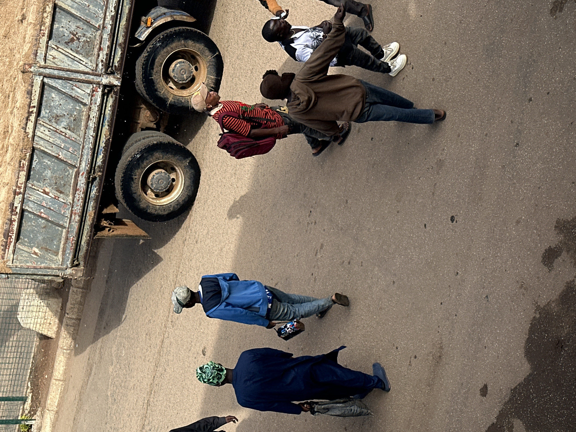 An overhead shot shows a group of people walking on a dusty road next to a large, dilapidated truck. The truck's cargo bed is filled with what appears to be sand or dirt. In the foreground, a person in a dark blue robe and green headscarf walks from left to right, carrying a large, dark bag in their left hand. To their right and slightly behind, another person in a blue hooded jacket and jeans walks in the same direction, carrying a small, patterned bag. Further to the right, a person in a red and black striped shirt and a tan baseball cap walks with a maroon backpack. Behind them, a person in a brown hooded jacket and jeans walks towards the right. To the far right, two other individuals are partially visible, one in a white and black patterned shirt and dark pants, and another in dark pants. The road is paved with asphalt and is speckled with dark spots, possibly oil or dirt. The environment appears to be an urban setting, potentially the city of Bukuru, Nigeria, given the context of the provided prompt. The lighting suggests it is daytime, with shadows cast by the truck and the people.