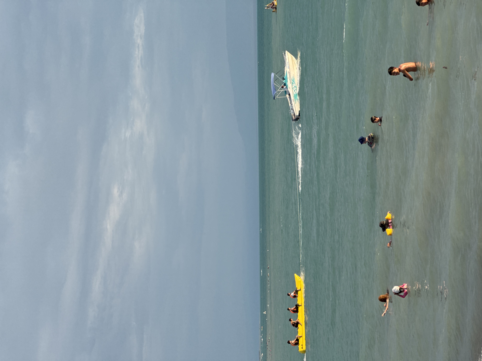 The image depicts a body of water with several people engaged in recreational activities. In the foreground, a young person wearing a pink swimsuit and a white hat is in the water. To their left, a person with long brown hair is partially submerged. To the right of the young person, another individual with a yellow flotation device and a dark hat is in the water. Further out in the water, several children are visible, including a young boy wearing a blue hat, another boy with dark hair, and a shirtless boy looking towards the camera. In the middle ground, a yellow banana boat is being pulled by a white speedboat, with four people seated on the banana boat, all wearing life vests. The speedboat has a blue canopy and a registration number "ABA 6181" visible. In the far distance, a hazy shoreline is visible with mountains beyond, under a cloudy sky. The water is a greenish-blue color. The scene appears to be a coastal or lakeside recreational area.