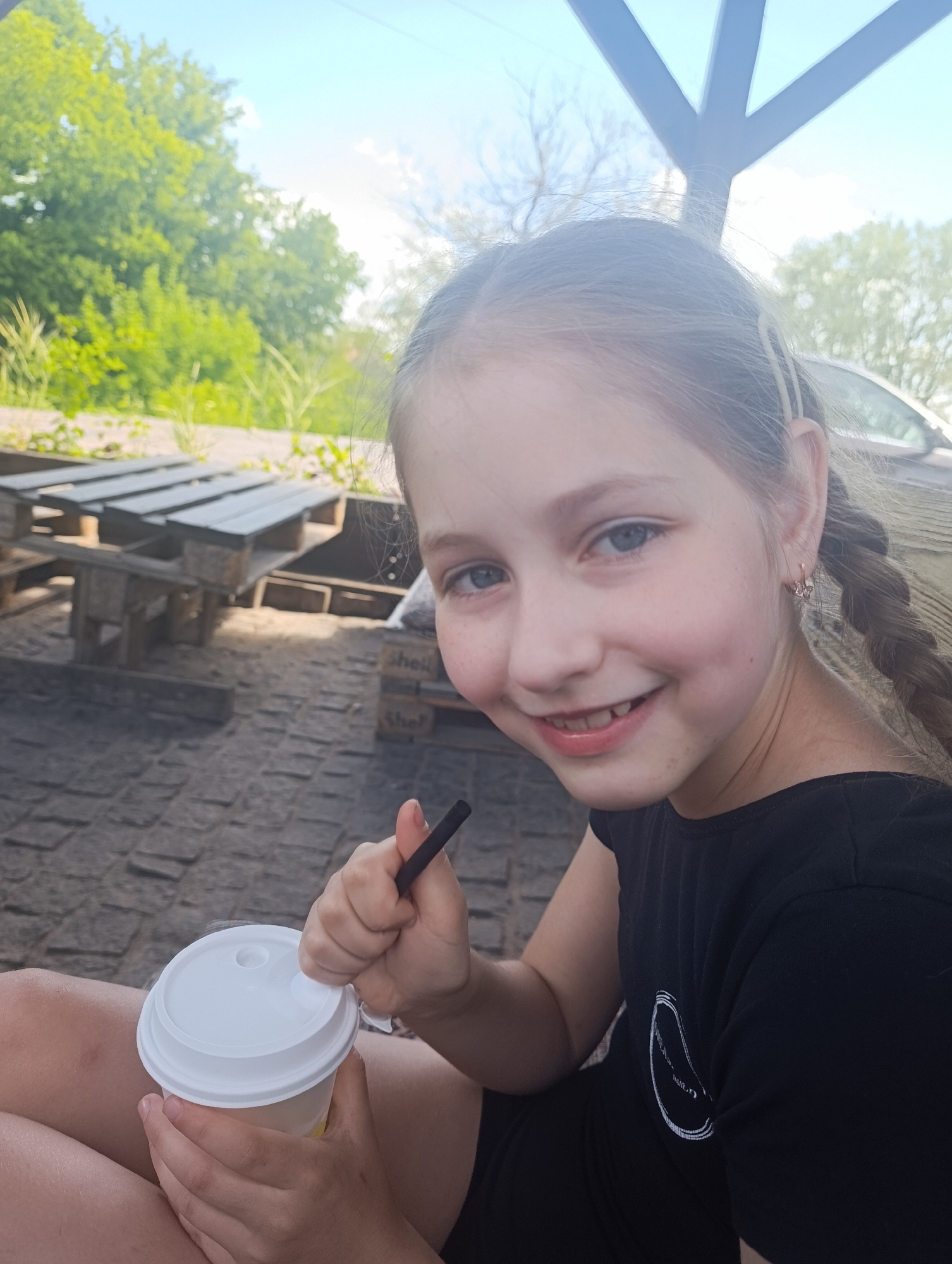 A young girl with blonde hair in braids and blue eyes smiles at the camera. She is wearing a black t-shirt and holding a white paper cup with a straw. The background shows an outdoor setting with wooden tables and chairs made of pallets, and lush green trees under a bright blue sky, suggesting it is daytime and the weather is pleasant. The girl appears happy and relaxed. The scene is likely a casual outdoor seating area, perhaps a cafe or park.