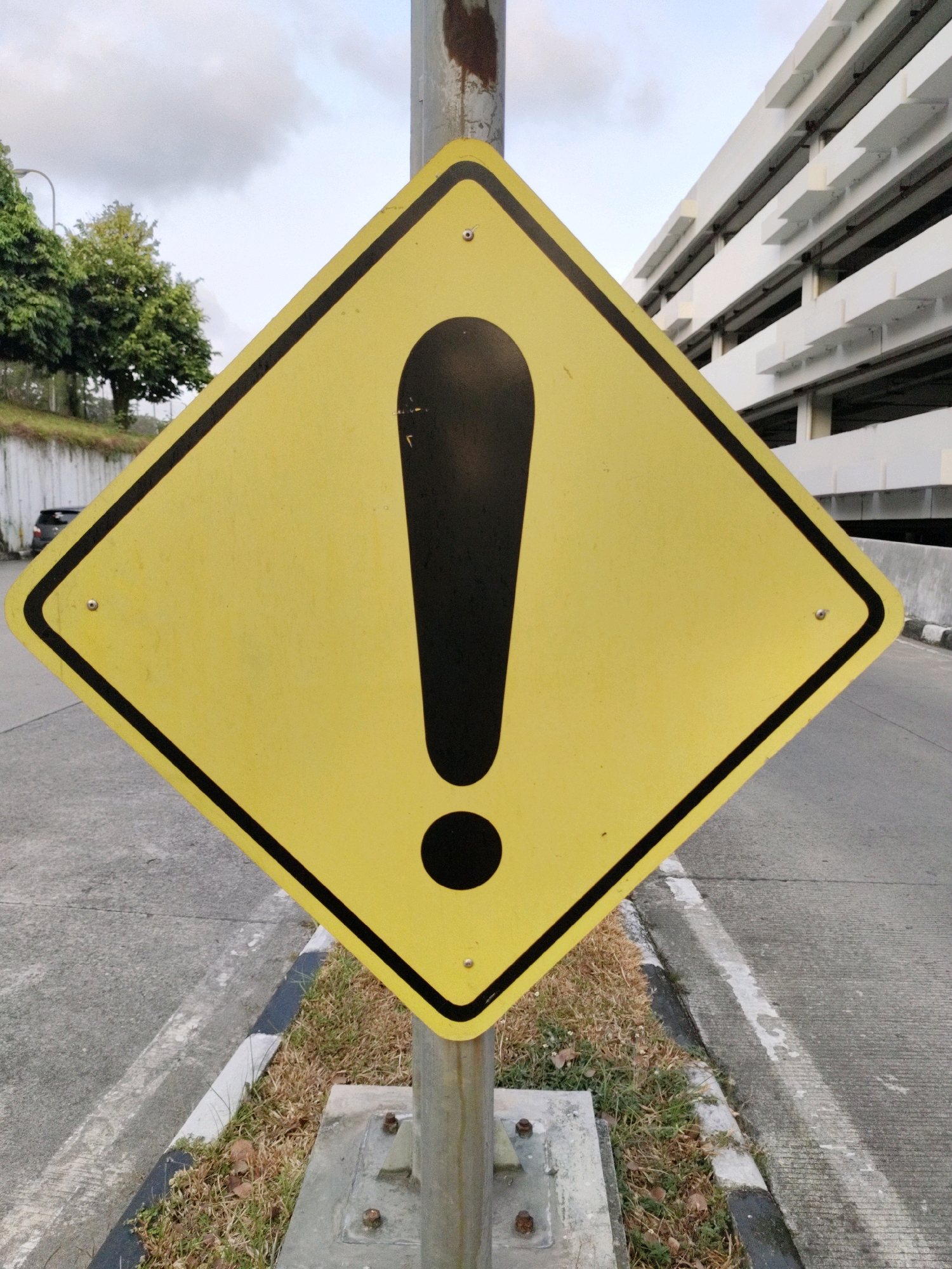 A diamond-shaped, yellow warning sign with a black border and a black exclamation mark is prominently displayed on a metal pole in Balikpapan, Indonesia. The sign is positioned at a slight angle, with its point facing upwards. In the background, a multi-story white parking garage dominates the right side of the frame. To the left, trees line a grassy embankment, and a street is visible with a car parked further down. The sign is situated near a paved road with a concrete curb. The sky is overcast.