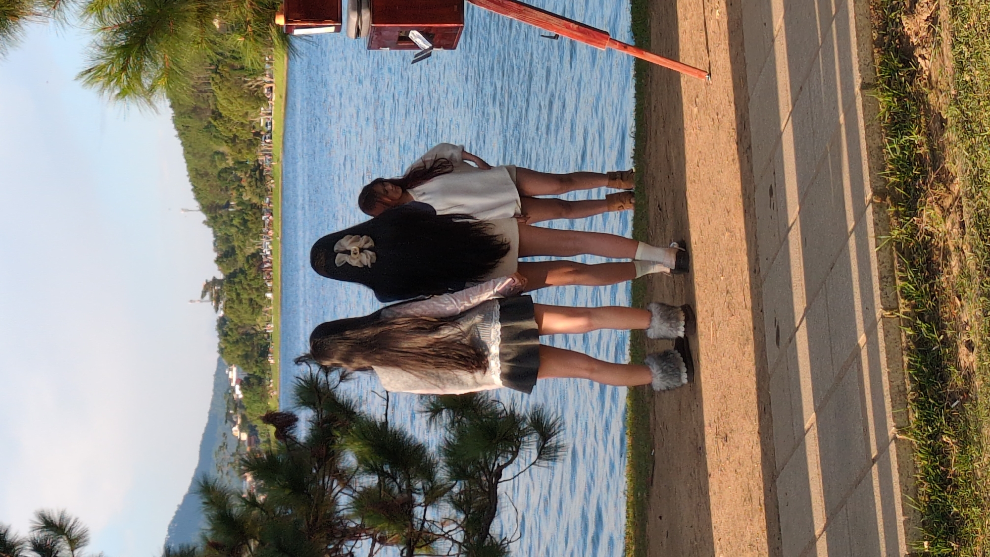 Three young women stand on the shore of Xuan Huong Lake in Da Lat, Vietnam, facing away from the camera. The woman on the left has long dark hair and wears a grey sweater over a white lacy top and a dark gray skirt. She has on fuzzy grey boot covers over dark shoes. The woman in the middle has very long dark hair adorned with a large, light-colored bow. She wears a light-colored short dress and white socks with dark shoes. The woman on the right has medium-length brown hair and wears a loose white shirt and dark shorts. She has on brown boots. Behind them is the blue water of the lake, with a tree-lined hill and buildings in the distance. To the right of the women, a vintage-style camera on a tripod is partially visible. The ground in the foreground is a sandy path next to a paved area with shadows cast by an unseen object.