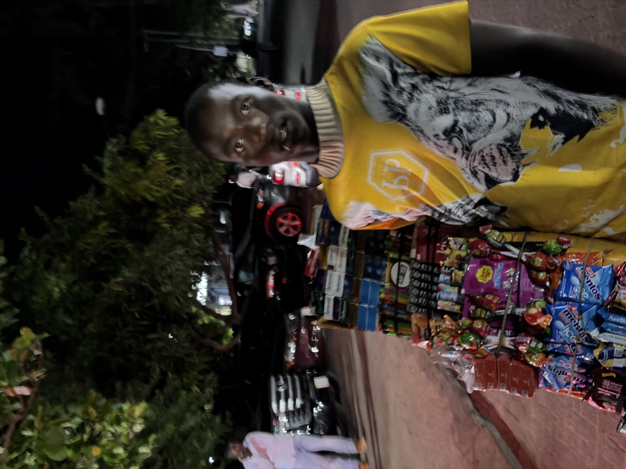 An outdoor photograph shows a man posing in front of a display of candy, gum, and cigarettes, presumably for sale at a roadside stand. The man, likely a vendor, has short dark hair and wears a yellow, white, and black patterned shirt. He is facing the camera with a slight smile.
Behind him, an assortment of items are arranged for sale: packets of Mentos, Chupa Chups, and other candies, along with packs of cigarettes and lighters.
In the blurred background, cars are visible, indicating a street scene, likely in Ikoyi, Nigeria.