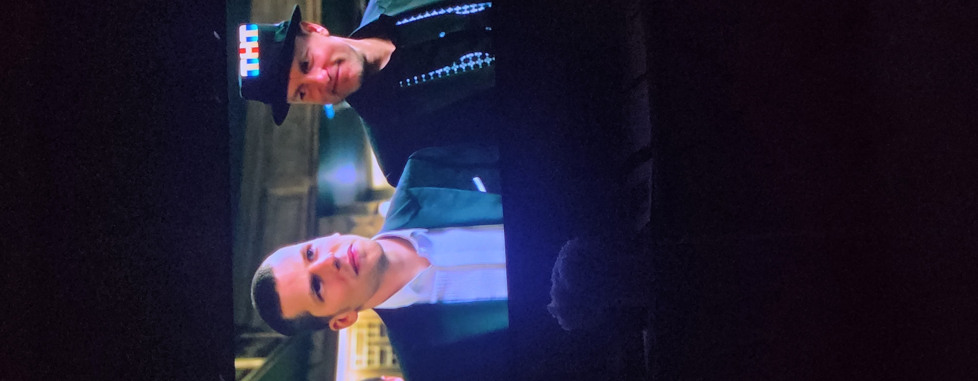 The image is a poorly lit, close-up, vertical shot of two men on a dark background. The man on the left is fair-skinned with a short haircut and is wearing a black suit jacket with a white collared shirt. He is looking slightly off to the right. The man on the right, who is only partially visible, is also fair-skinned and is wearing a black hat with the letters "THT" lit up in a bright, colorful pattern on the front. He is wearing a black shirt and suspenders with a patterned design. He appears to be looking to his right. The lighting in the image is dark, and the details of the background are obscured. The scene is not identifiable as being in Kazan, Russia.