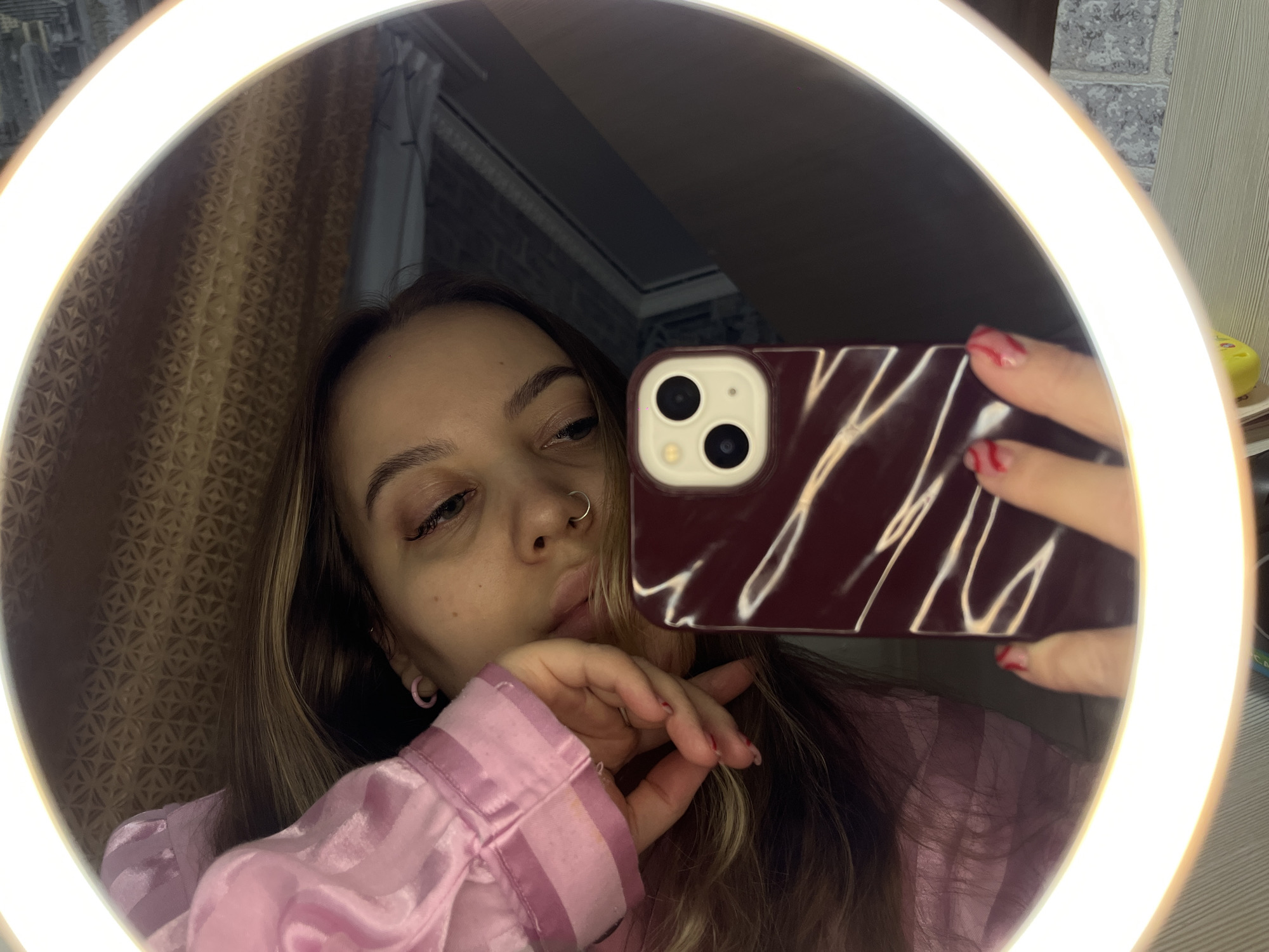 A person is taking a selfie in a round mirror lit by a ring light. The individual has light brown hair with blonde highlights and is wearing a pink satin-like shirt. They have a nose ring and pink earrings. They are holding a burgundy phone case with a light-colored phone that has two visible camera lenses. The background includes a patterned curtain and a wall, possibly in a residential setting. The image location is stated as Novopokrovskaya, Russia, but this detail cannot be independently verified from the visual content of the image.