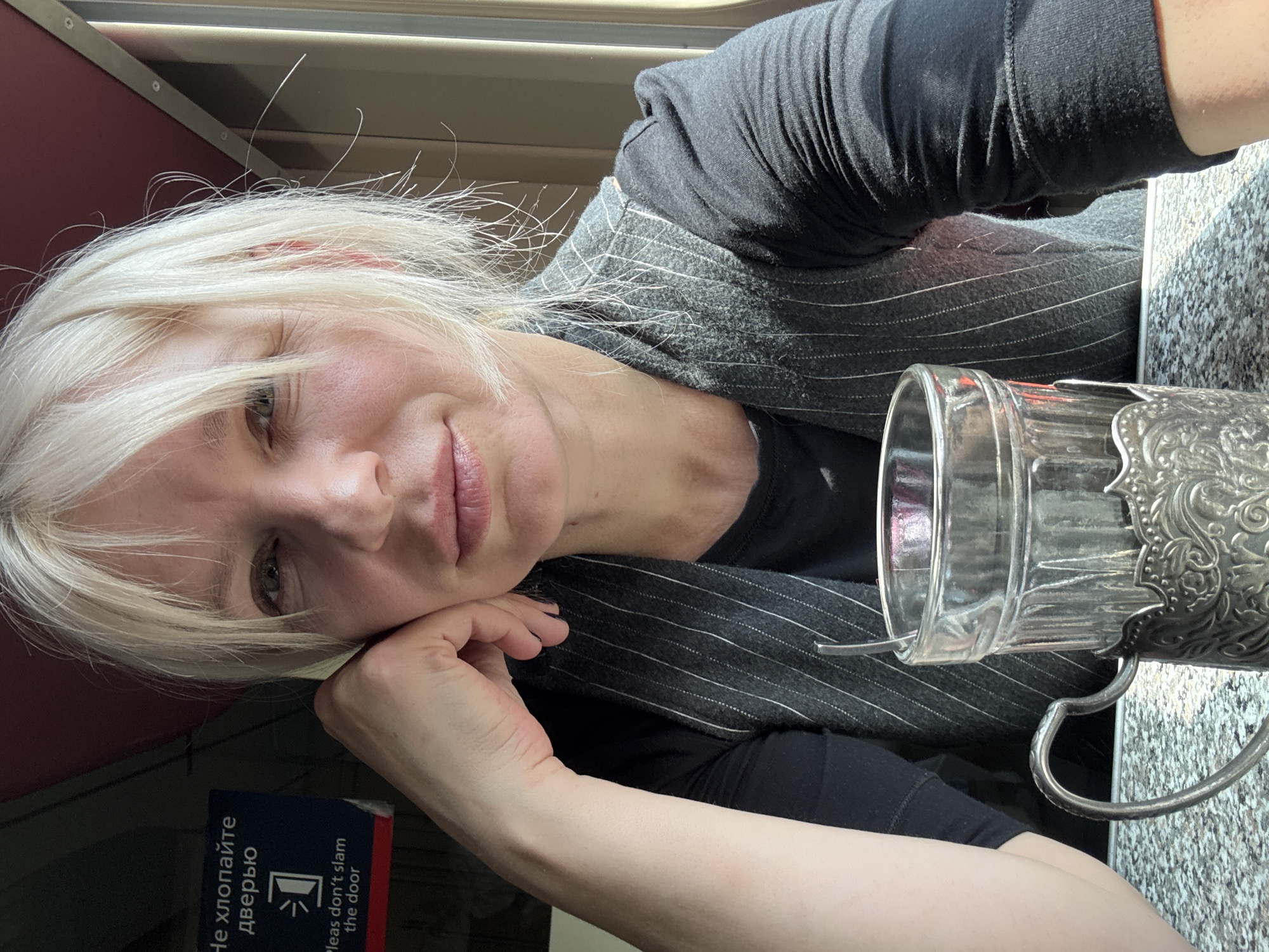 A woman with blonde hair and fair skin is pictured in a selfie on a train. She is wearing a dark grey vest over a black long-sleeved shirt. Her right hand is resting on her chin, and she is looking towards the camera with a neutral expression. In the foreground, a glass with a silver ornate holder is visible, with a spoon inside. A sign in Russian and English is partially visible on the left, with the text "Не хлопайте дверью" and "Please don't slam the door". The background suggests the interior of a train compartment, with a window to the right showing an outdoor scene. The overall lighting indicates it is daytime. The location is identified as Shirokaya Balka, Russia.