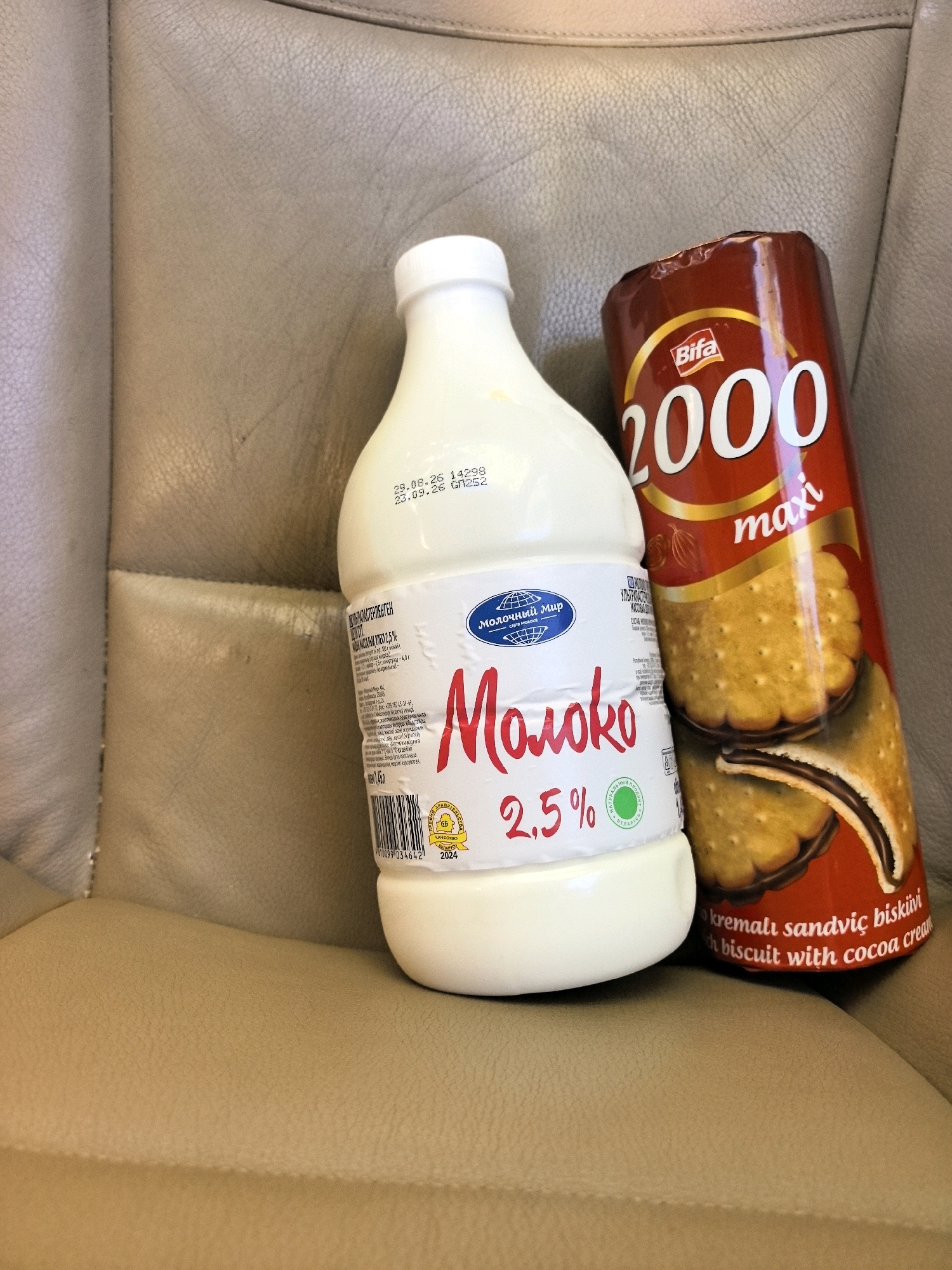 In Astrashitskagaradokski rural council, Belarus, a bottle of "Moloko" (milk) with a 2.5% fat content and a package of Bifa "2000 maxi" biscuits with cream and cocoa filling are placed on a tan-colored leather surface, likely a car seat. The milk bottle is white with a white cap and features a label with Russian text and a blue logo depicting a globe. The biscuits are in a red and gold cylindrical package. The image is taken from a slightly elevated angle, focusing on the two products. The background is a blurred continuation of the leather material.