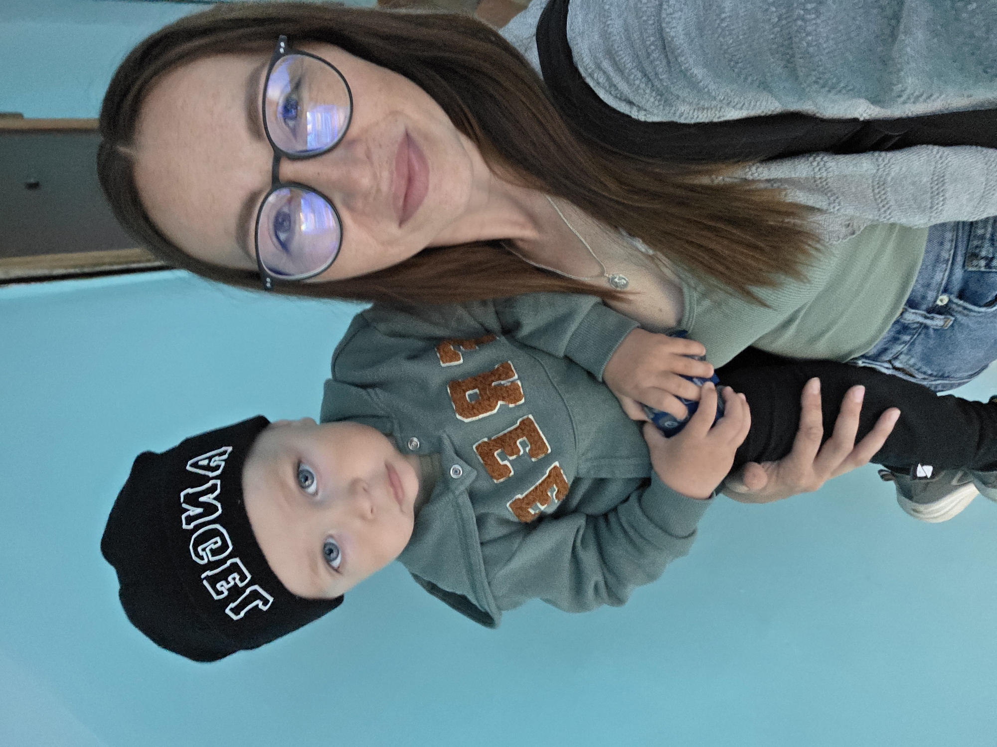 A woman with long brown hair and glasses is holding a young child. The child is wearing a black beanie with white lettering that spells "ANGEL" backwards, and a green hoodie with brown lettering that appears to spell "REEB". They are standing in front of a bright blue wall, possibly in a public space or residential area. The woman is wearing a light green top and a gray cardigan. The child is holding a small blue object. The overall impression is of a casual, everyday moment. The location is not specified in the image.
