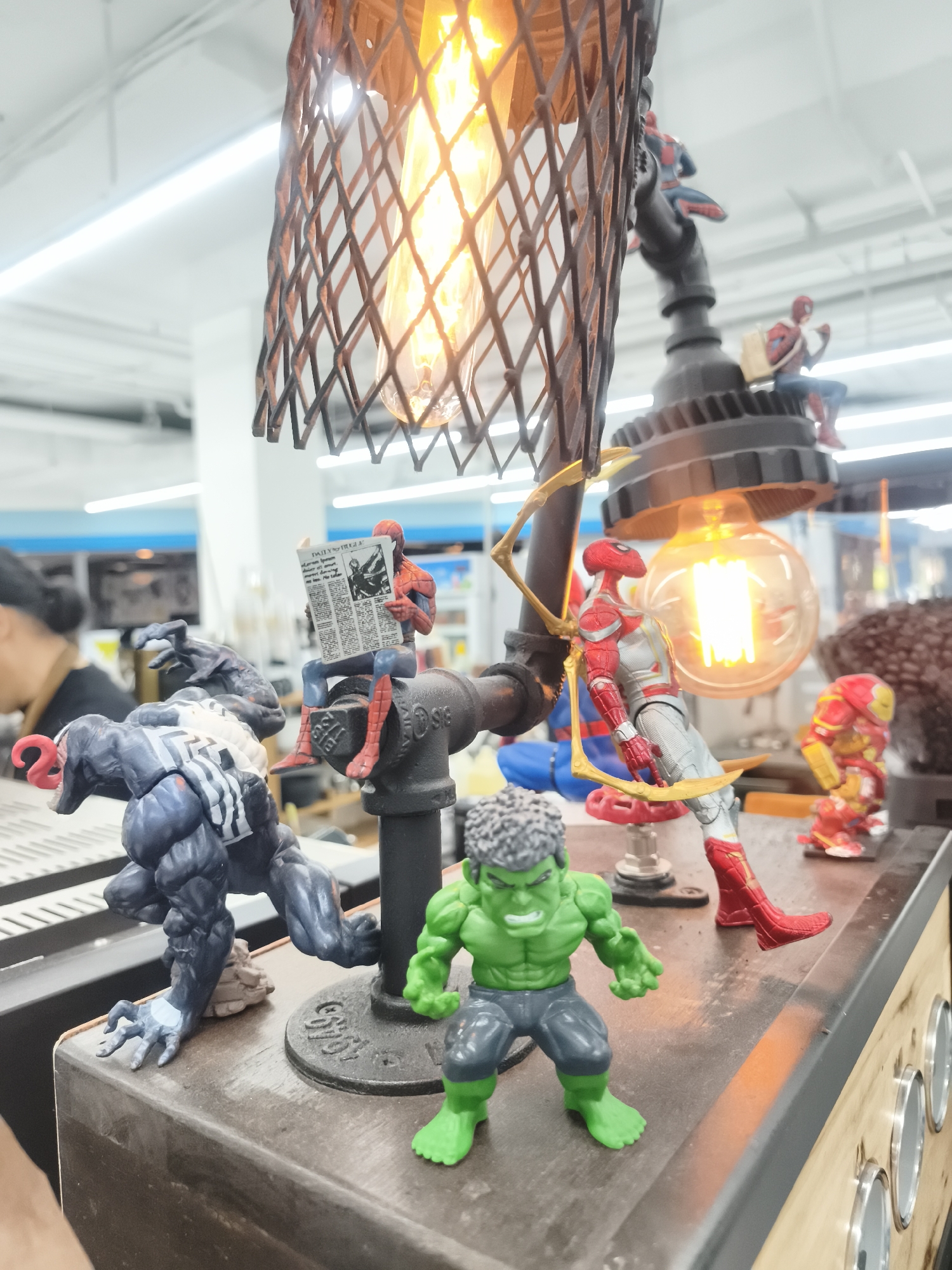 The image shows a lamp constructed from metal pipes and fittings, adorned with Marvel superhero figurines. The lamp has two exposed light bulbs: one within a wire cage and the other exposed. The figurines include multiple Spider-Man figures in various poses, Venom, Hulk, and Iron Man. One Spider-Man figure is seated reading a newspaper. The lamp appears to be placed on a wooden surface with metal circular accents along its side. In the blurred background, there's a person with dark hair and a brightly lit interior space. The location is บ้านบางรักษ์, Thailand.