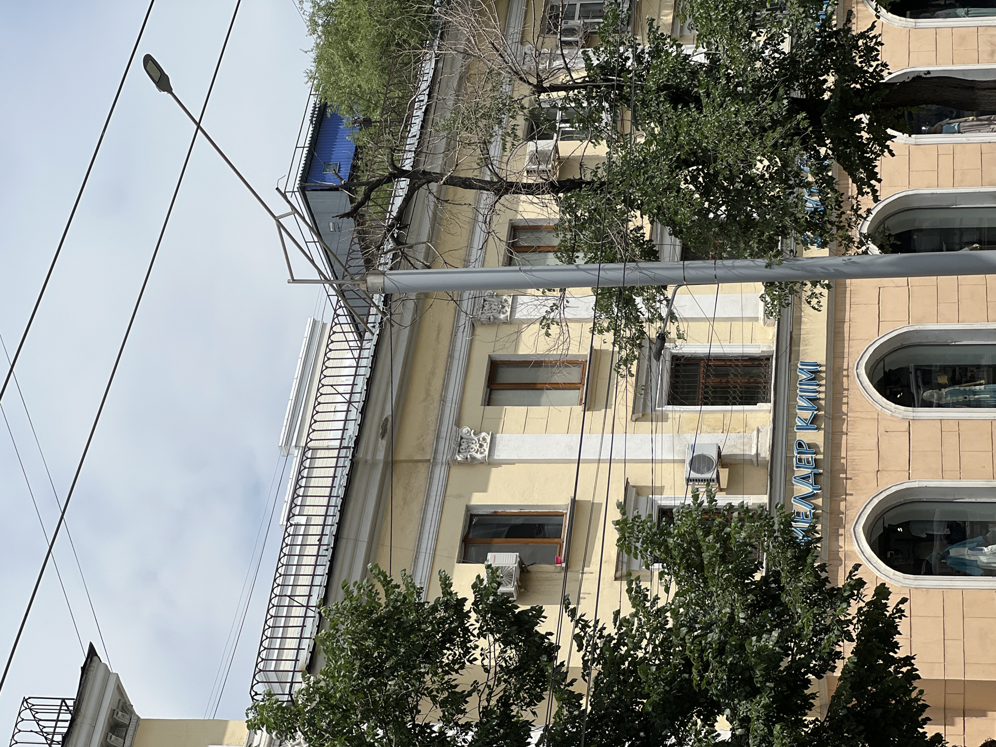 The image shows a section of a yellow, multi-story building on a street in Almaty, Kazakhstan. The building has arched storefronts on the ground floor, one of which displays mannequins. Above the storefronts, there are windows on the second and third floors, some with visible air conditioning units. A sign with blue, backlit lettering, reading "МЕДЕЕР КИИМ", is mounted above the arched windows. Trees with green foliage partially obscure the facade. Overhead power lines and a street light are visible against a cloudy sky.