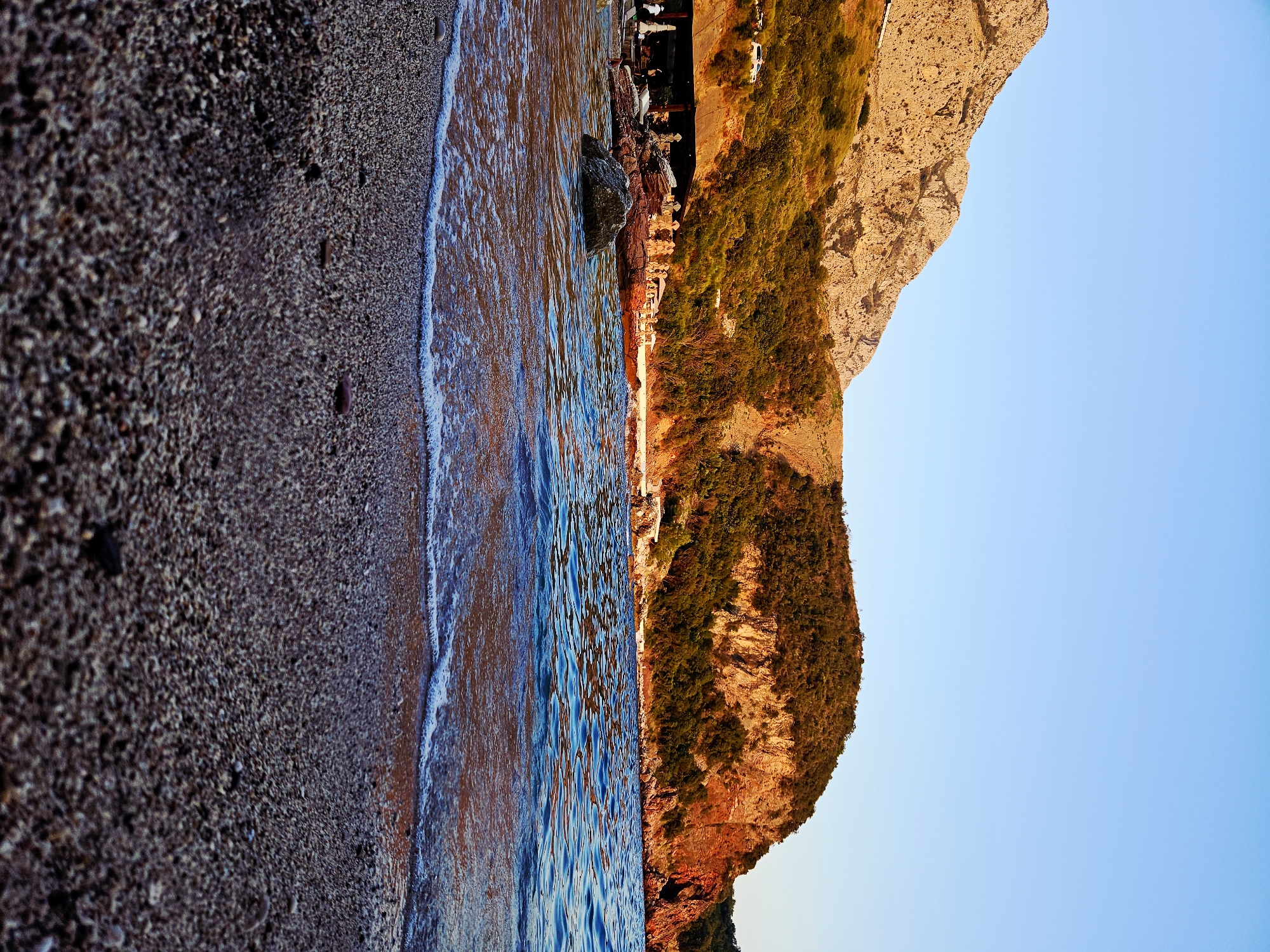 The image depicts a picturesque coastal scene at dusk, likely located near Sveti Stefan, Montenegro. The foreground is dominated by a pebble beach, wet from the receding tide, with small waves gently lapping at the shore. The water itself is a deep blue, reflecting the warm hues of the setting sun.
In the midground, a rocky outcrop juts into the sea, adorned with sparse vegetation. Behind this, a covered, open-air structure resembling a restaurant or beach bar is visible, with tables and chairs set up. Several people are present, some appearing to be staff and others patrons, enjoying the evening ambiance. A few white umbrellas are also set up on the beach.
The background features a dramatic hillside covered in dense, green foliage, transitioning to rugged, rocky terrain further up. The imposing mountains, bathed in the warm, golden light of the setting sun, create a striking backdrop. The sky above is a clear, pale blue, indicative of the fading daylight. The overall impression is one of serene natural beauty and a relaxed, vacation-like atmosphere.