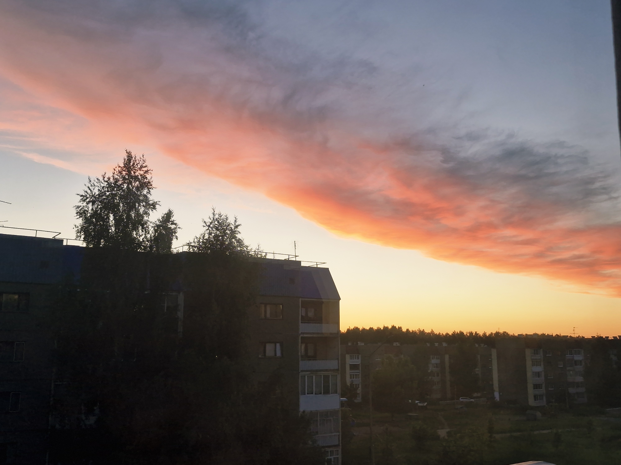 The image captures an urban or suburban landscape at either sunrise or sunset, dominated by a vibrant and colorful sky. A long, horizontal cloud formation stretches across the upper half of the frame, illuminated in brilliant shades of pink, orange, and red, gradually fading into lighter blue and gray tones towards the top. The lower portion of the sky near the horizon glows with a warm golden-orange light.
In the foreground, a dark, multi-story residential building with a blue roof, balconies, and visible windows is positioned on the left, partially obscured by the dark silhouettes of several trees. Antennas are visible on its rooftop. Further in the mid-ground, other blocky apartment buildings are visible, interspersed among clusters of trees. A dense treeline marks the horizon, separating the urban structures from the colorful sky. Faint outlines of cars can be seen on the ground between the buildings. The low angle of the sun casts much of the foreground and ground level into shadow, while accentuating the dramatic colors of the clouds. No people or specific activities are depicted, and there is no visible text. The scene conveys a sense of serene natural beauty during twilight.