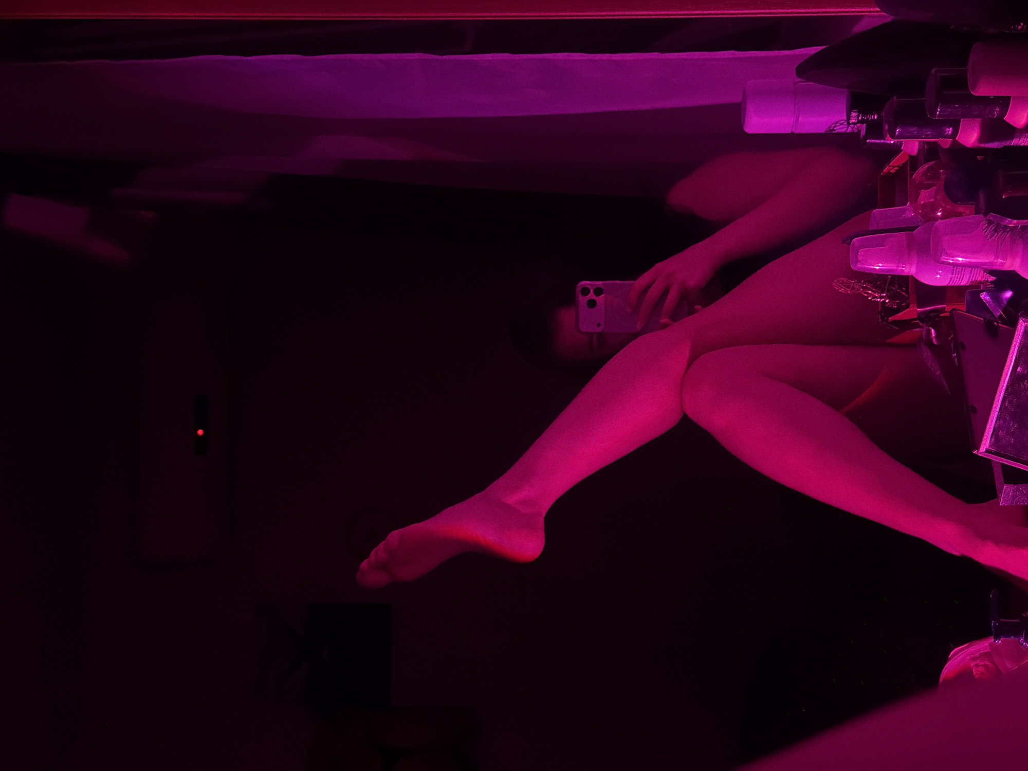 A person in a room is taking a selfie with a phone. The person's bare legs are visible, as well as the phone they are holding. Various bottles and containers are arranged on a surface next to the person. The room is dark and lit with pink lighting. A small electronic device with a red indicator light is mounted on the wall. The image does not depict or infer Moscow, Russia.