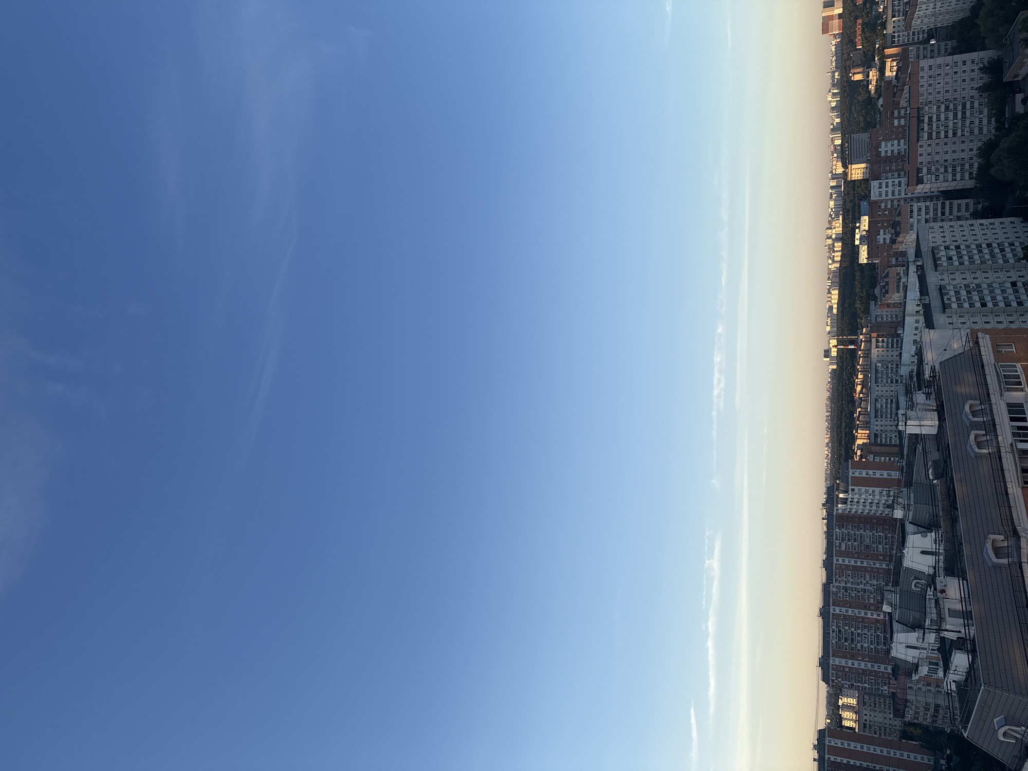 This is a vertical, eye-level, wide shot of a city skyline against a clear blue sky. The image is split into two distinct halves: the left side is dominated by the vast expanse of the sky, transitioning from a deep blue at the top to a lighter shade near the horizon. Thin, wispy clouds are scattered across the sky, particularly noticeable in the upper left quadrant. The right side of the image showcases a dense urban landscape, with numerous multi-story apartment buildings stretching as far as the eye can see. The buildings are a mix of architectural styles, with many featuring light-colored facades and rows of windows. Some buildings are taller than others, creating a varied silhouette against the sky. The tops of some buildings reveal roofing details, including vents and what appear to be dormer windows. Trees are visible interspersed between the buildings, suggesting the presence of parks or tree-lined streets within the city. The sunlight casts a warm glow on the buildings, indicating it is either early morning or late afternoon. The overall impression is of a sprawling, lived-in city under a peaceful sky.