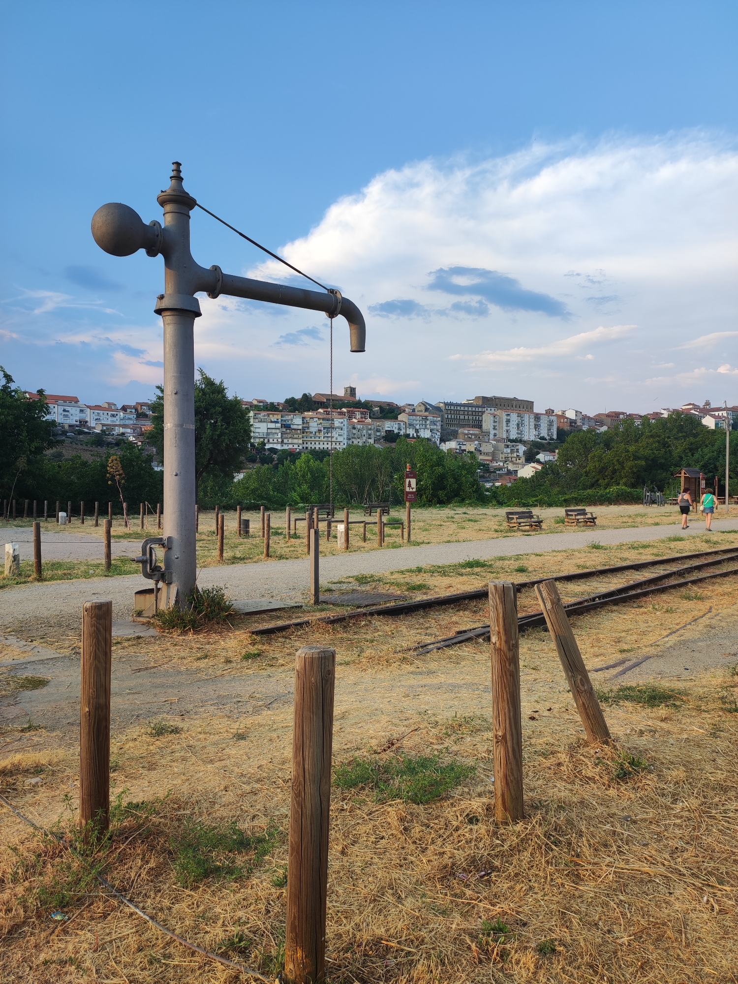 In Béjar, Spain, a vintage water pump, constructed from weathered metal and featuring a large spherical handle and a downward-curving spout, stands prominently in the foreground. The pump is situated in a dry, grassy area, with a light-colored gravel path running alongside it. Beyond the path, a set of train tracks, rusted and weathered, stretch into the distance. Several wooden posts, some with reflective markers, line the area, suggesting a maintained pathway or boundary. In the middle ground, wooden benches are placed along another gravel path. Further back, a verdant line of trees separates the park area from a hillside town, characterized by numerous buildings with red-tiled roofs and white facades. The town is built on a gentle slope, with taller structures visible towards the crest. The sky above is a clear blue, punctuated by scattered white clouds, indicating fair weather. Two women are visible in the middle distance, walking along a path, their figures small against the broader landscape. The overall scene suggests a public park or historical site, likely a former industrial area repurposed for recreational use, with the train tracks hinting at a past connection to transportation or industry. The light suggests late afternoon or early evening.