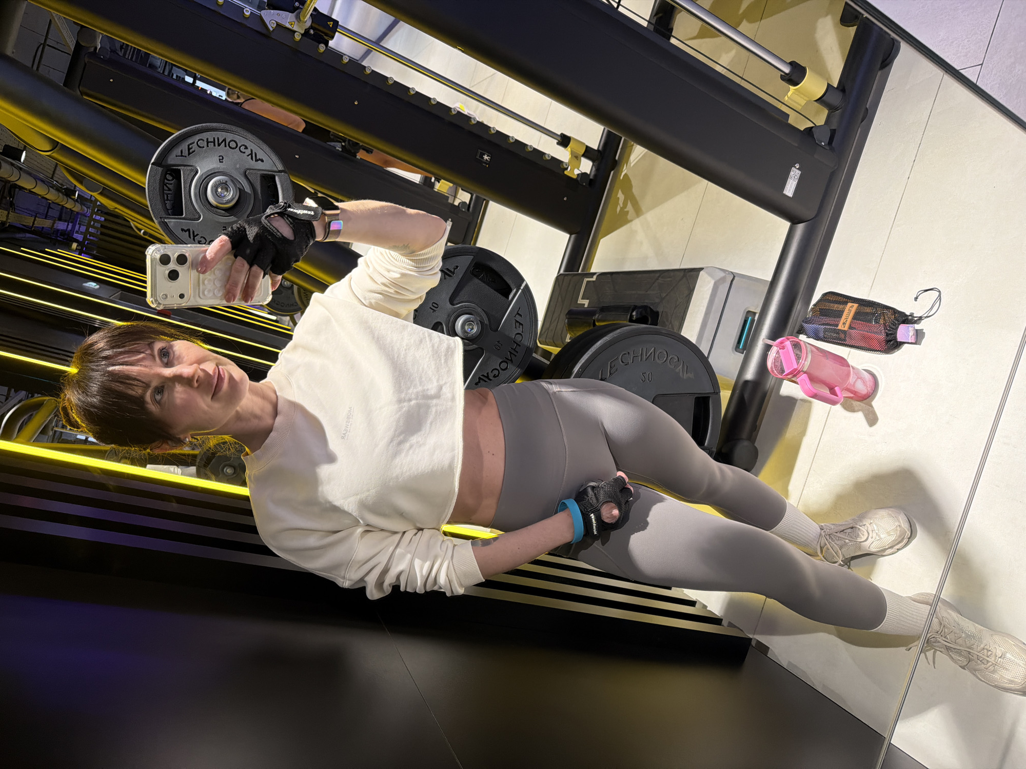 The image captures a woman taking a selfie in a modern gym in Perm, Russia. The woman, appearing to be in her 30s or 40s with brown hair and bangs, is looking directly at the camera with a neutral expression. She is positioned on a piece of gym equipment, possibly a leg press machine, which features dark surfaces and prominent yellow LED strip lighting.
She is wearing a white cropped long-sleeved sweatshirt, gray leggings, white socks, and light-colored athletic shoes. On her hands, she has black fingerless workout gloves, with "Everlast" visible on her right glove and "gymbeam" on her left. A light-colored smartwatch or fitness tracker is on her right wrist, and a blue wristband adorns her left. She holds a white iPhone with a clear case in her left hand, using it to take the photograph.
In the background, several large black weight plates are visible, some branded "TECHNOGYM" and marked with numbers such as "50", "2", "10", and "20". Nearby on the floor are a pink water bottle and a small black mesh bag containing colorful resistance bands. The gym interior features a combination of dark, sleek equipment and light-colored tiled flooring with reflective surfaces, contributing to a clean and contemporary aesthetic.