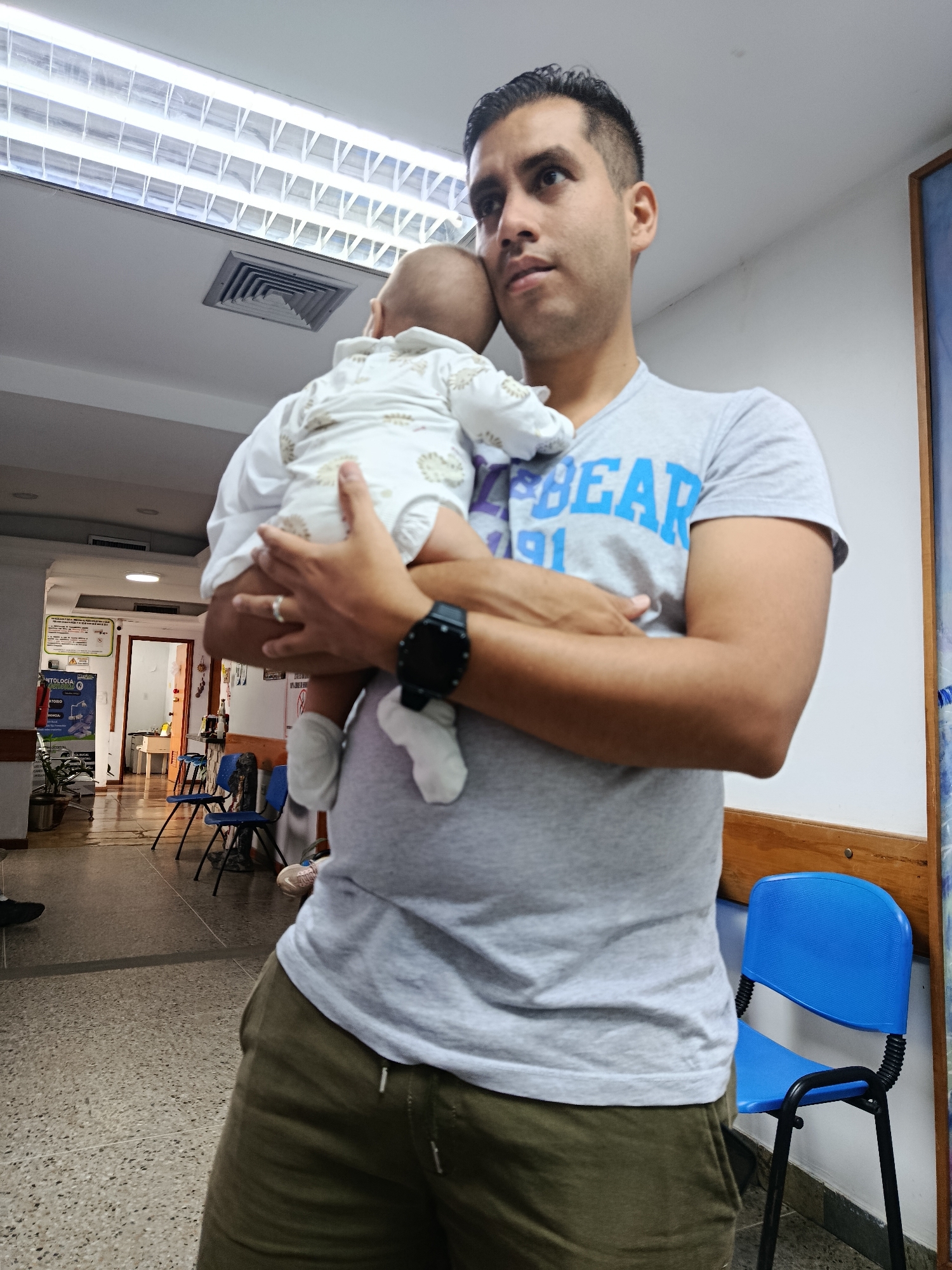In the interior of what appears to be a medical waiting area in Juan Griego, Venezuela, a man is holding a baby. The man, with dark hair and a grey t-shirt with blue lettering, cradles the baby, who is wearing a white onesie with a subtle pattern. The baby is facing away from the camera, resting its head on the man's shoulder. The man is looking off to the side with a pensive expression.
The environment shows fluorescent lighting on the ceiling and a ventilation grate. In the background, a hallway with several blue chairs is visible, along with potted plants and informational posters on the wall. The floor is tiled. The overall scene suggests a moment of quiet waiting, possibly in a clinic or hospital.