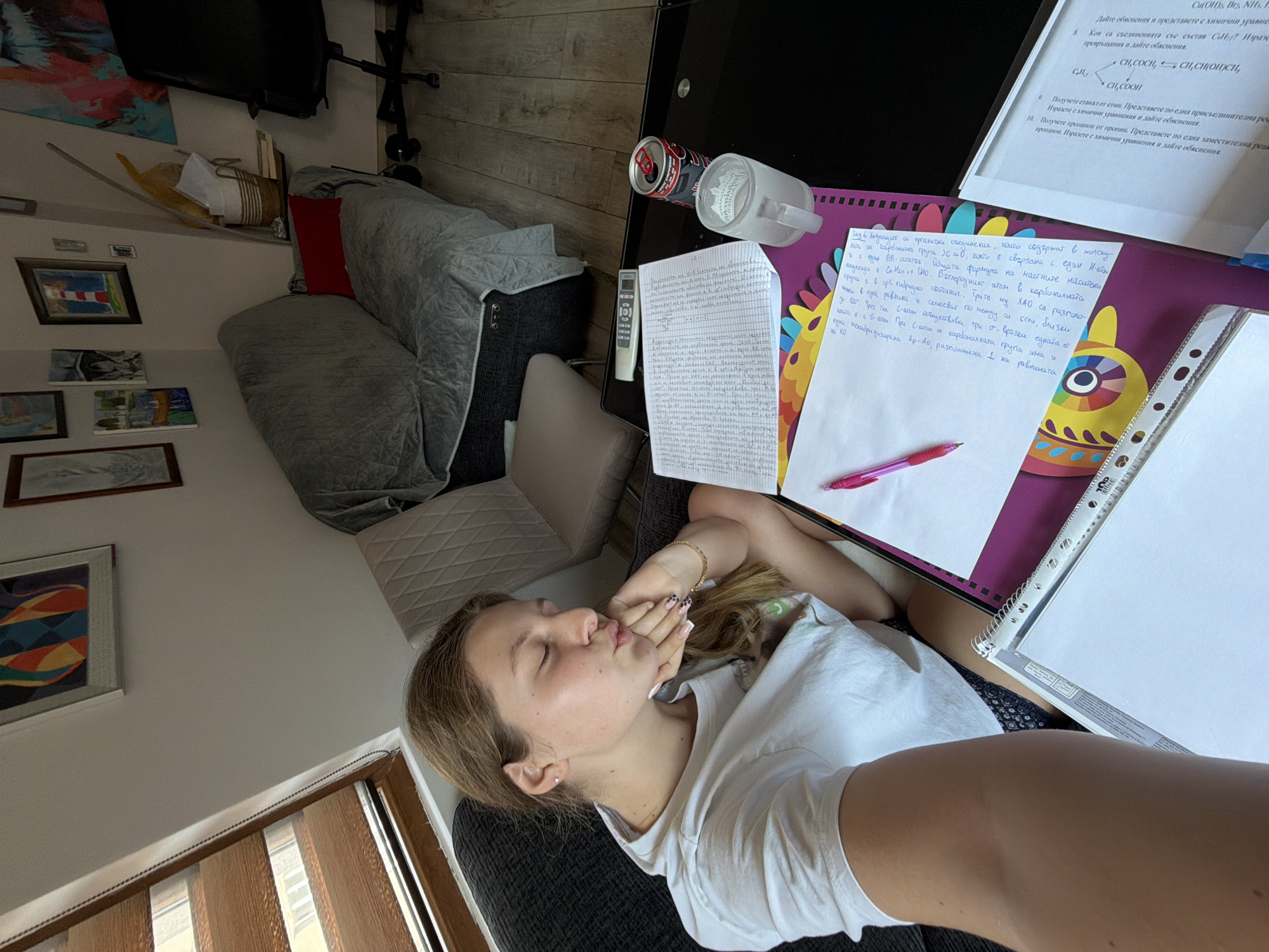 In the city of Burgas, Bulgaria, a young woman with light brown hair is pictured from the chest up, sitting on a dark couch. She is wearing a white t-shirt and has her eyes closed, with her hand resting on her chin and her lips pursed, as if blowing a kiss. She has manicured nails with a patterned design. In front of her, on a black table, is a notebook, a pen, a can of Coca-Cola, and a glass mug. The room appears to be a living area, with a sofa covered with a grey quilted blanket, another chair, and artwork on the wall. The lighting suggests it is daytime, with light filtering in from a window with wooden blinds.