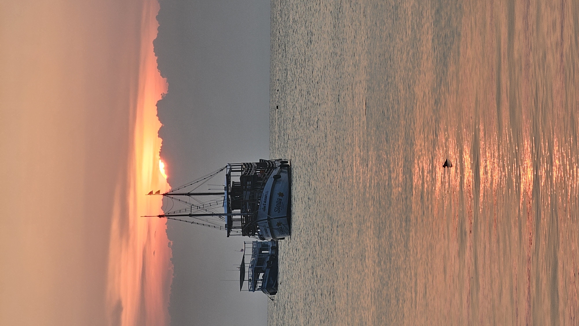 The image shows a sunset scene in Tambon Ko Tao, Thailand. In the water are two boats. The boat closest to the viewer has the word "Stella" written on the side. The sun is setting behind clouds on the left side of the frame. The water reflects the orange color of the sky. A bird appears to be flying over the water.