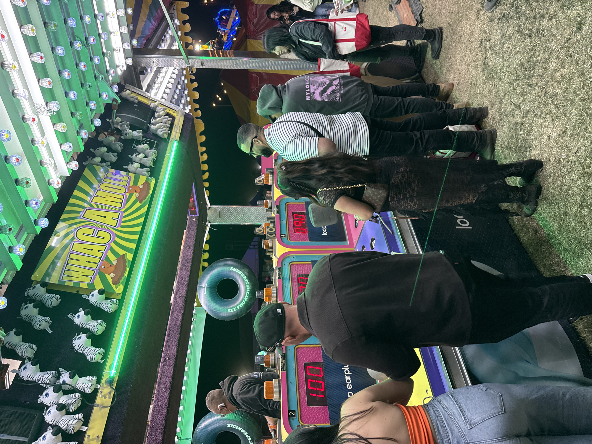 Here is a forensic description of the image:
**Scene:** A nighttime image of a "Whac-A-Mole" carnival game being played by a group of people. The game is situated within a carnival booth, featuring a colorful display and various components. The location is assessed to be a public event likely in the city of Thermal, California, United States of America.
**People:** The image features multiple individuals of varying heights and builds standing shoulder to shoulder playing the Whac-A-Mole game. Their attires vary from casual wear to more dressed-up clothing.
**Objects:** The key objects within the image include:
* The "Whac-A-Mole" game structure itself, with its prominent signage and components such as the game's control panel and prize display area above.
* Individual display screens showing the score of each player.
* Plush zebra toys in the prize area
* "SEE YOU AT THE SPEAKERS" labeled buoys hanging off to the side.
**Actions:** The people are actively engaged in playing the game, hitting the moles with a mallet.
**Interactions:** There are interactions between players and the game as they compete for a high score, and amongst each other.
**Environment:** The image captures a dynamic environment with carnival lighting. The ground appears to be grass or a similar surface.