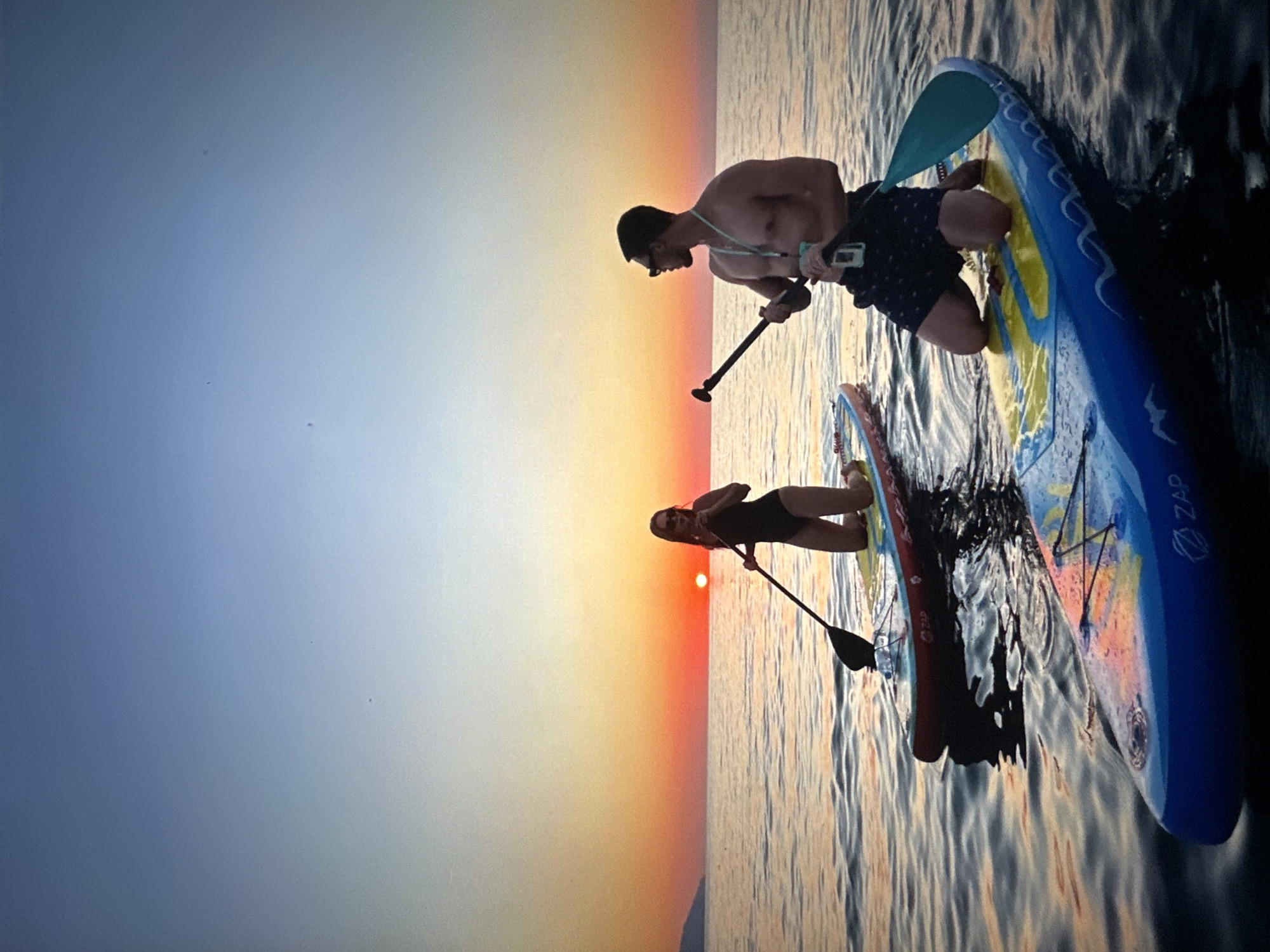 The media file shows two people on paddleboards in the water during a sunset. The person in the foreground is a man wearing dark shorts and sunglasses, kneeling on a blue and yellow paddleboard. He is holding a paddle and appears to be looking at something in his hand. Behind him, a woman in a black swimsuit is on another paddleboard, also holding a paddle. She is looking towards the sunset. The sun is low in the sky, casting a warm orange glow that transitions to yellow and then to a pale blue in the upper sky. The water is calm with gentle ripples reflecting the colors of the sunset. The overall mood is serene and peaceful, capturing a moment of outdoor recreation at dusk. Visible text on the paddleboard in the foreground reads "ZAP".