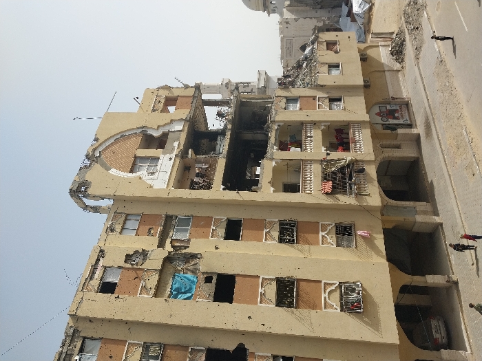 The image displays a multi-story building in Khan Yunis, Palestinian Territory, exhibiting extensive structural damage under a clear, light blue sky. The central building, rendered in a light brown facade with darker brown accent panels, shows widespread destruction, particularly on its upper floors and right side. Numerous windows are missing or shattered, revealing dark, debris-filled interiors and exposed rebar. Bullet or shrapnel impacts are visible across the facade. An ornate arched architectural feature on the building's upper right is partially destroyed. Balconies on the lower-middle floors, featuring white railings, have clothes hanging from them. The ground floor consists of arched openings, one of which shelters a parked silver sedan on the left.
A paved street runs horizontally in the foreground, interspersed with patches of dirt and debris. On the left side of the street, three individuals are walking away from the camera. On the right, a single individual stands facing the damaged building. To the right of the main structure, significant piles of rubble and further damaged buildings are visible, including the partially obscured white dome of a structure, possibly a mosque, in the background.
