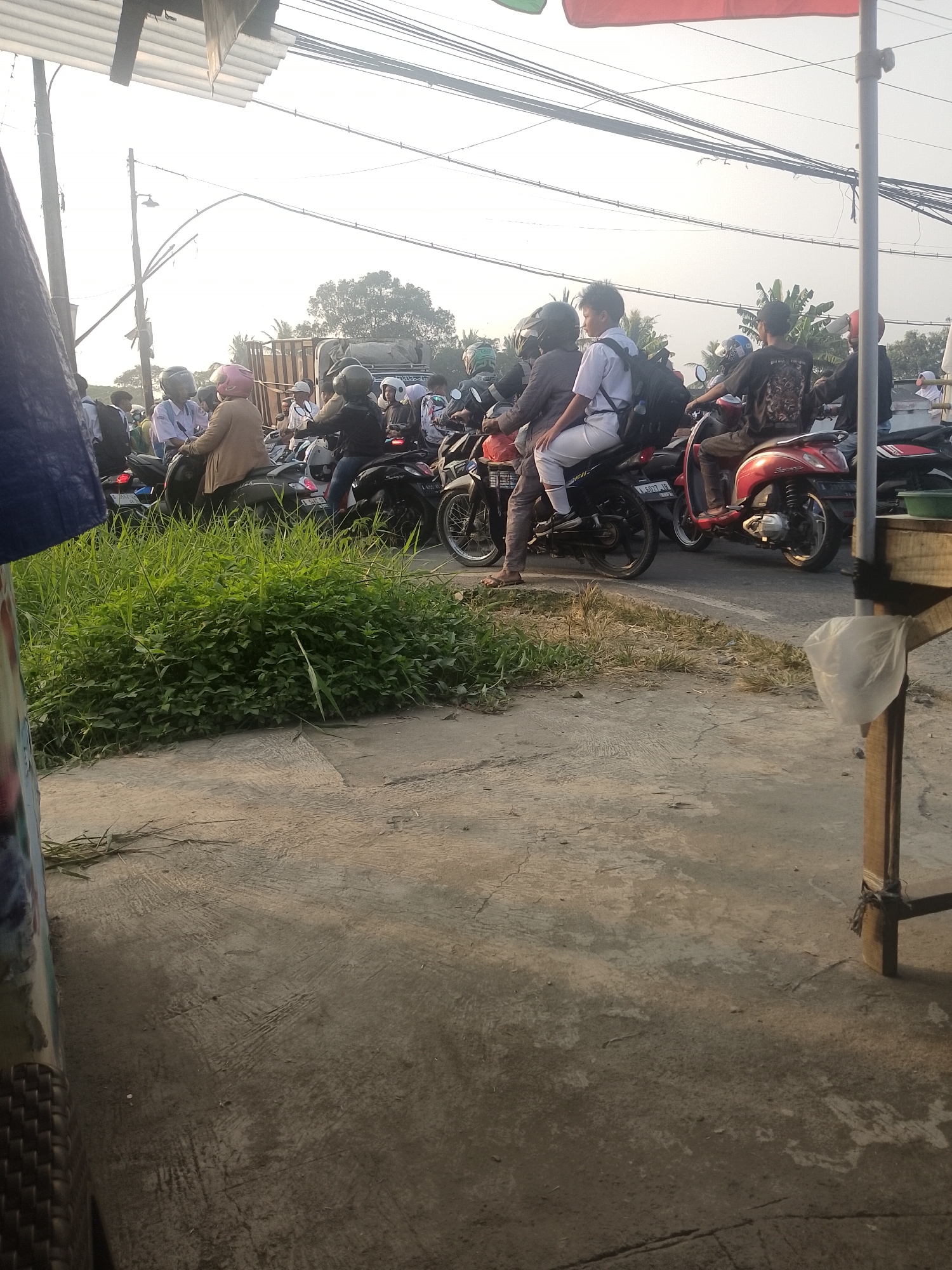 The image depicts a street scene in Ciruas, Indonesia, likely during daylight hours, given the bright sky. The foreground is dominated by a concrete surface with patches of dry grass and some green vegetation on the left. A wooden structure with a white plastic bag tied to it stands on the right. In the mid-ground, a traffic jam of motorcycles and their riders is visible. Several individuals are seated on motorcycles, some wearing helmets. One motorcycle has a red body. Further back, a truck is partially visible, along with more motorcycles and people. Overhead, a network of electrical wires crisscrosses the sky, indicating an urban or semi-urban environment. The overall impression is one of a busy, possibly slow-moving traffic situation on a street.