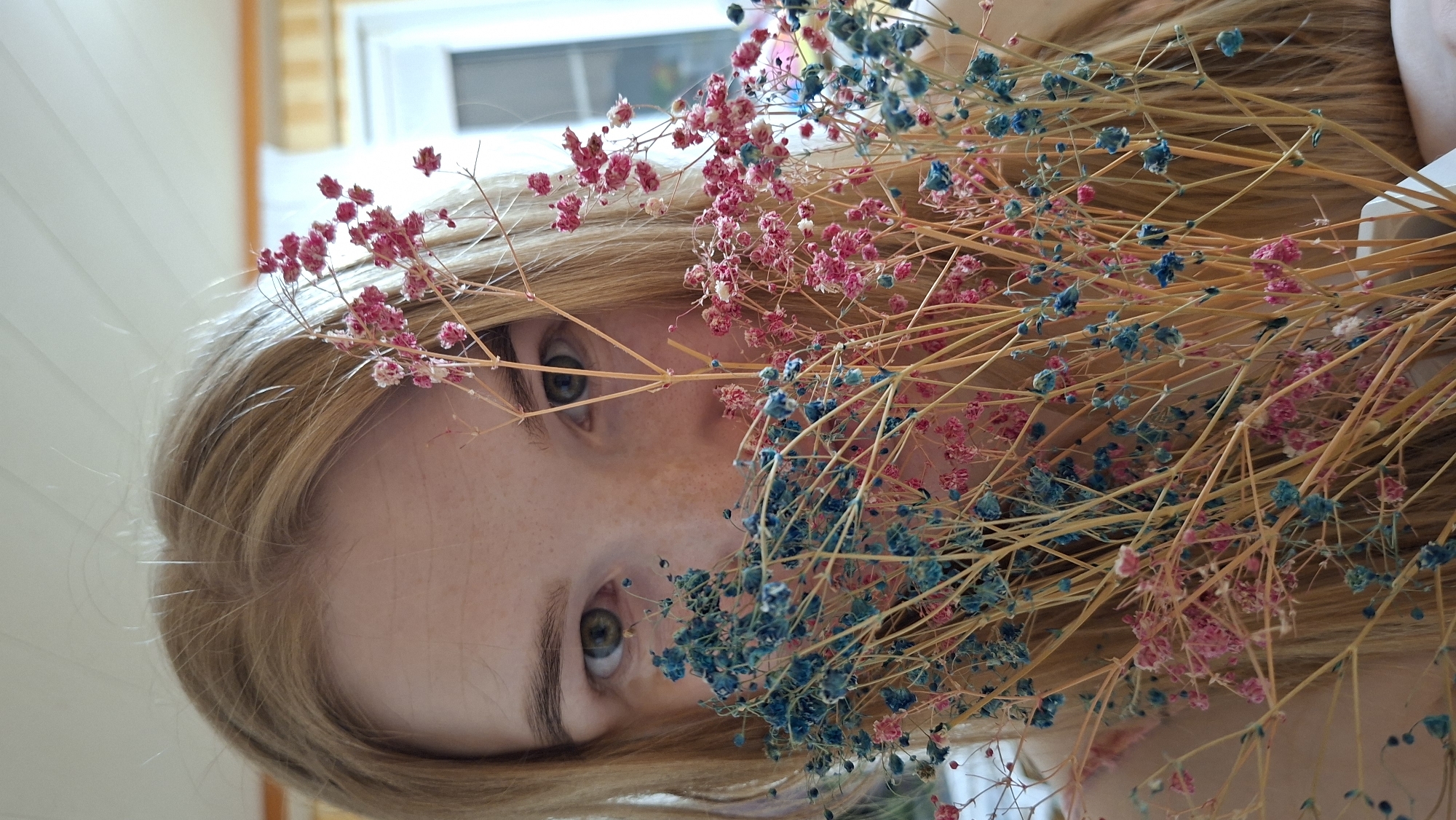 A close-up image shows a person with light blonde hair and visible freckles on their face, looking directly at the viewer. The face is partially obscured by a bunch of dried baby's breath flowers, dyed in pink and blue hues. The person's eyes are green. The background shows a blurred interior with vertical white paneling and a window with a white frame and a window pane. The location in the city of Большой Букор, Russia is not determinable from this image.