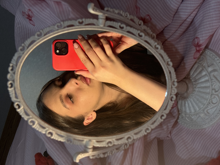 An image depicts a person taking a selfie using a red smartphone, reflected in an oval, ornate tabletop mirror. The scene is presented from a slightly rotated perspective, with the person's reflection appearing horizontally across the mirror.
The reflection shows a young individual, likely female, with long, dark brown hair that falls around their head. Their face is visible, with the left eye slightly lowered or looking off-camera, a delicate nose, and glossy, slightly parted lips. They are wearing subtle eye makeup, possibly eyeshadow and mascara, and their lips appear to have a warm-toned gloss. The lighting highlights the bridge of their nose and the fullness of their lips.
A hand, with neatly manicured fingernails painted with a natural-looking, possibly French tip, holds a bright red smartphone with a visible triple-lens camera setup, characteristic of an iPhone Pro model. The phone is positioned in front of the person's face, indicating they are capturing their own image in the mirror.
The mirror itself has an elaborate, off-white or cream-colored frame with intricate, carved or molded details around its perimeter. The base of the mirror, also ornate and matching the frame, is partially visible both within and outside the reflection.
The background outside the mirror's frame features a soft, light-colored fabric, possibly a bedsheet or blanket, patterned with thin stripes and scattered small red or pink bows. The overall lighting is soft and warm, casting gentle shadows and creating a somewhat intimate atmosphere. No text is visible in the image.