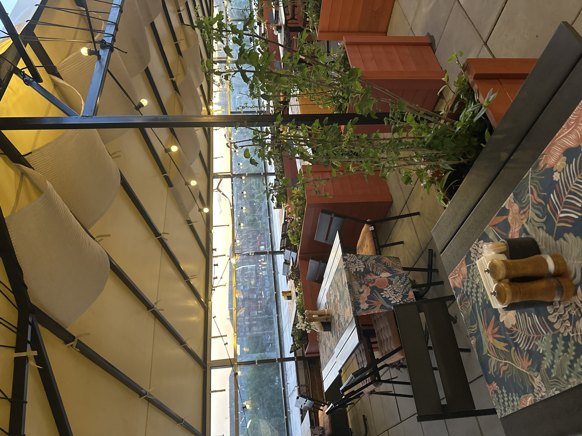 The image depicts an outdoor dining area, likely a restaurant or cafe, in what appears to be Perm, Russia, given the visible urban landscape through the glass walls. The area is partially covered by a structured awning with fabric draping, adorned with string lights that create a warm ambiance. There are wooden planters containing greenery, separating dining spaces. Tables are set with decorative floral tablecloths, condiment containers and chairs. Through the glass walls, buildings and urban features can be seen, suggesting an urban environment. No people are clearly visible, but the setting implies it's prepared for customers.