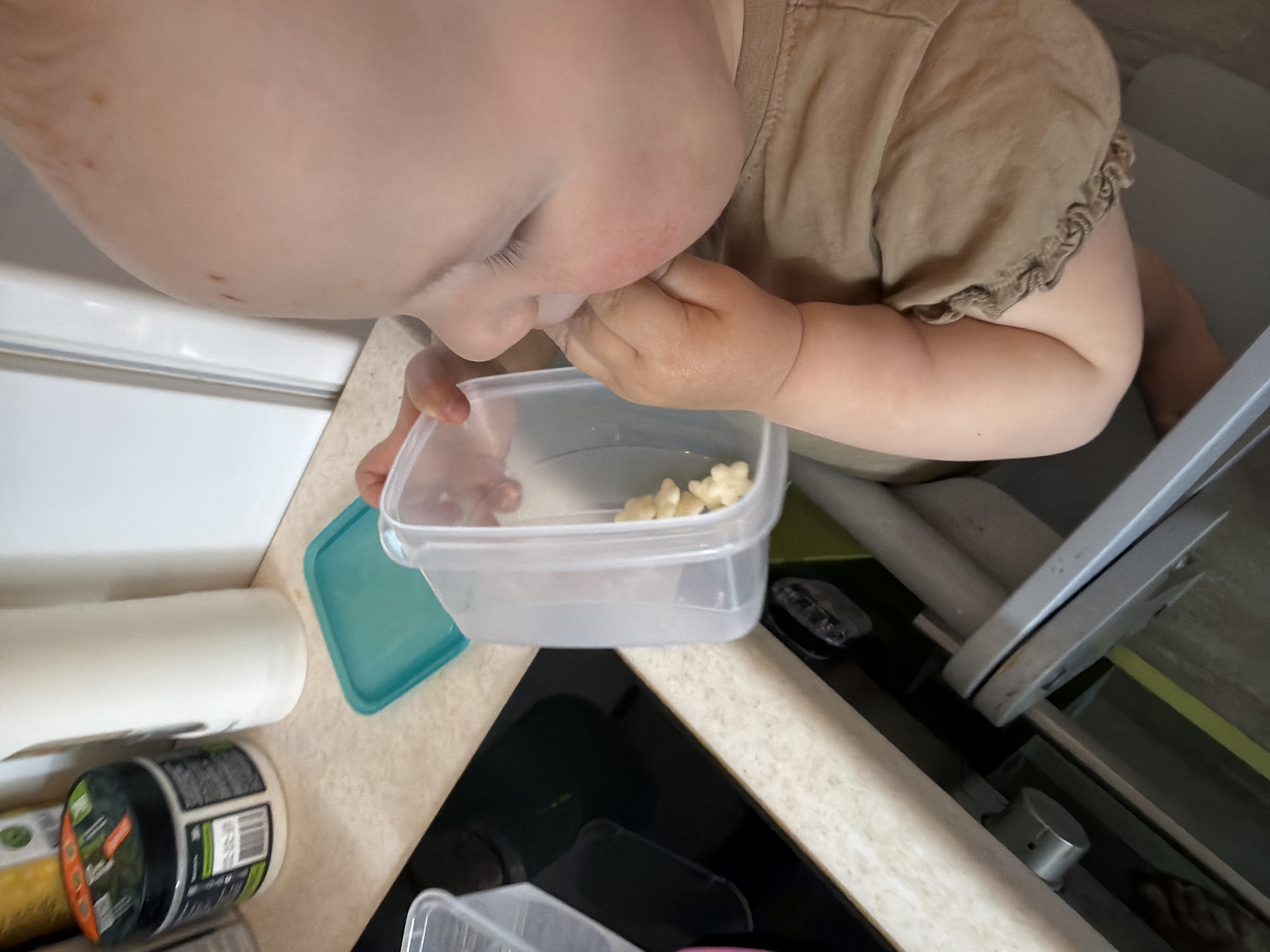 A baby, wearing a beige shirt with frilled sleeves, is seated in a high chair and eating puffed grain snacks from a clear plastic container with a blue lid. The container is on a countertop next to a roll of paper towels and a jar. The high chair is located next to a stove. The location is likely a kitchen, but the specific city, Chaikovsky, Russia, cannot be determined from this image.