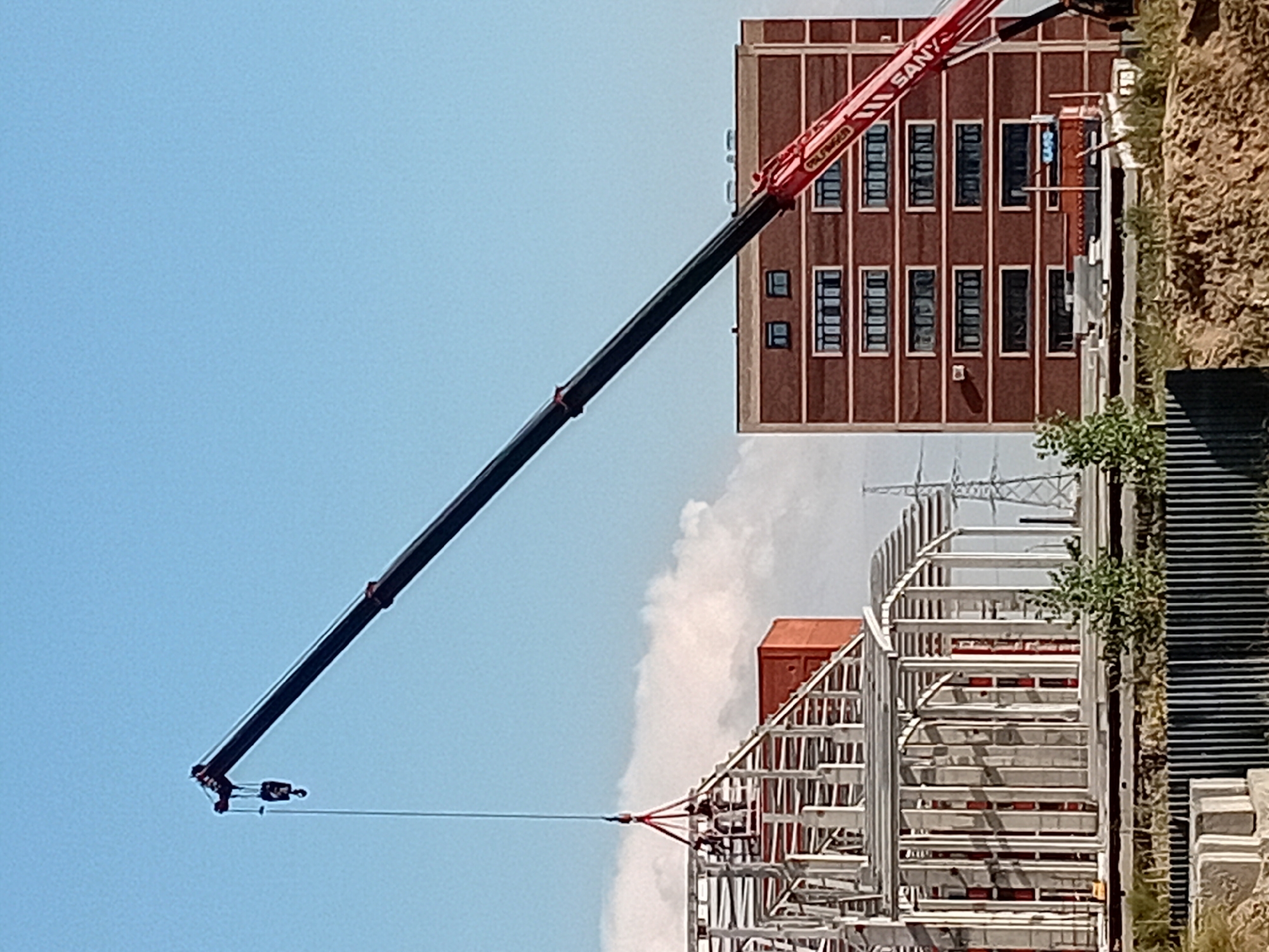 In Semey, Kazakhstan, a construction site is depicted with a large crane extending into a clear blue sky. The crane's boom is angled upwards, with its hook suspended in the air. Below, a building with a steel frame is under construction, showcasing its skeletal structure. Adjacent to this, a multi-story brick building stands completed, featuring rows of windows. A power pylon is visible in the background, hinting at the surrounding urban or industrial landscape. The foreground shows a rough, earthen embankment and some construction materials. Two individuals are visible on the steel frame of the building, engaged in construction activities. The scene captures a moment of progress and development within the city.