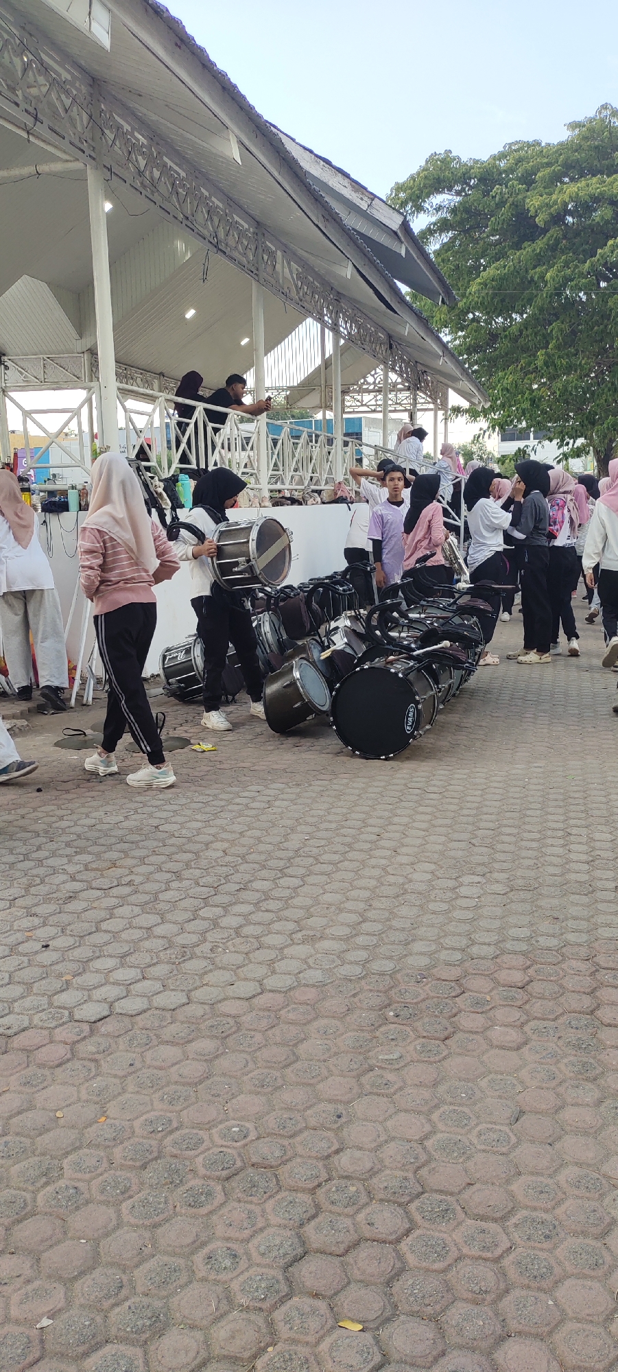 A group of young people, some wearing hijabs, are gathered around a collection of marching band drums on a hexagonal paver ground. The scene appears to be outdoors, possibly at an event or practice. A white, open-air structure with a corrugated roof is visible in the background, suggesting it might be a stage or covered seating area. Trees are also present in the background. Some individuals are standing and observing, while others are interacting with the drums. One person is holding a drum and stick. The overall atmosphere suggests a pre-performance or post-performance setting. The location is identified as Lhokseumawe, Indonesia.