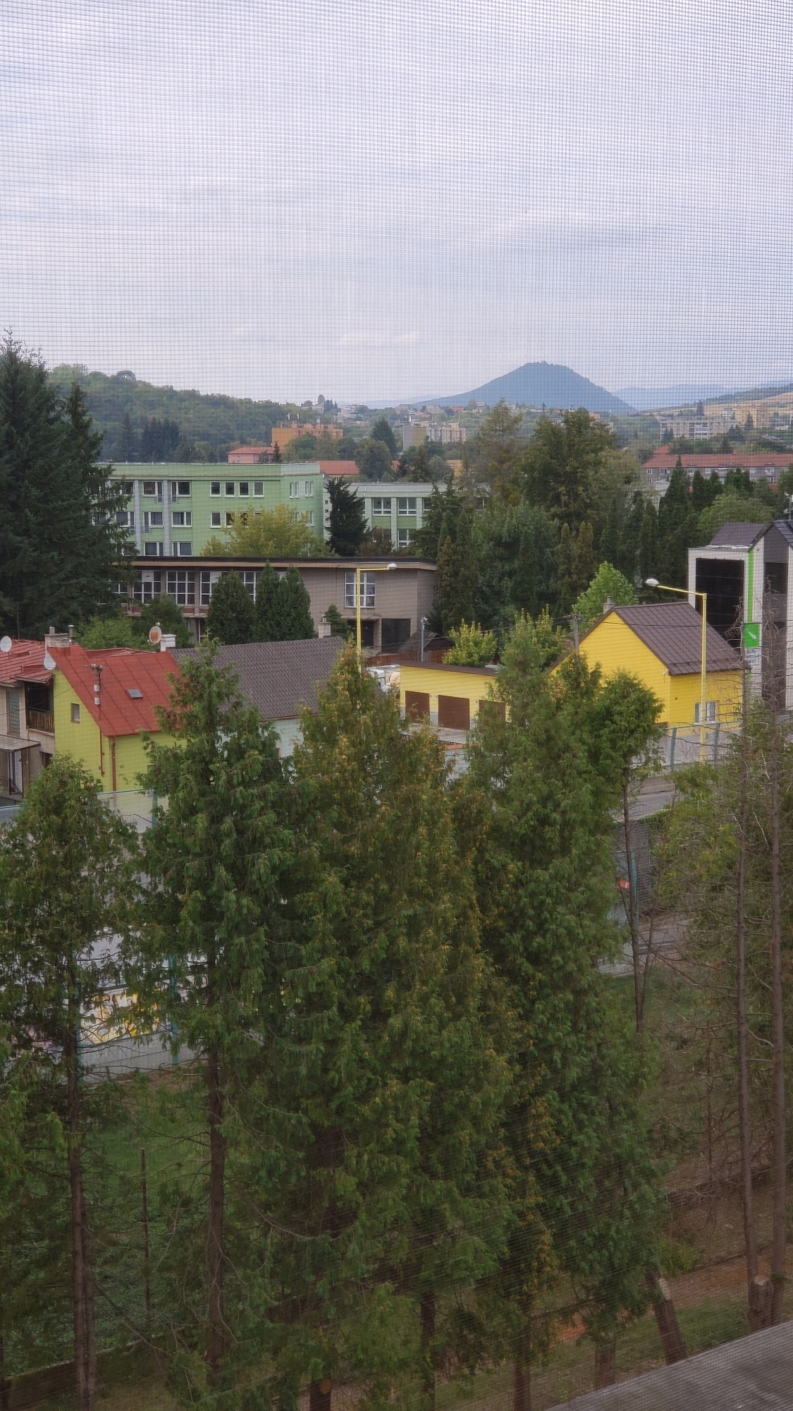 The image captures a view of a residential area in Prešov, Slovakia, looking out from behind a screen. In the foreground, several evergreen trees with dense foliage dominate the scene, obscuring much of the lower portion of the view. Behind the trees, a cluster of houses and other buildings are visible.
To the left, a yellow house with a red roof is prominent, with an adjacent structure partly visible. Further back, a row of taller buildings, appearing to be apartment blocks or institutional buildings, are painted in light green and have multiple windows. One building has a dark roof with a prominent chimney. To the right of these green buildings, a lighter-colored, two-story building with large windows is visible.
In the middle ground, a bright yellow building with a brown roof stands out, possibly a garage or workshop, with three large doors. A single yellow lamppost is positioned in front of it. Further back, more houses and buildings are scattered amongst trees, with some appearing to have red tile roofs.
The background of the image features a rolling, tree-covered hillside and a distinct, cone-shaped mountain with a visible structure on its peak, which is characteristic of the landscape surrounding Prešov. The sky is overcast with a grid pattern, likely from the screen through which the photo was taken. The overall impression is of a residential neighborhood on the outskirts of the city, with natural elements and urban development intermingled.