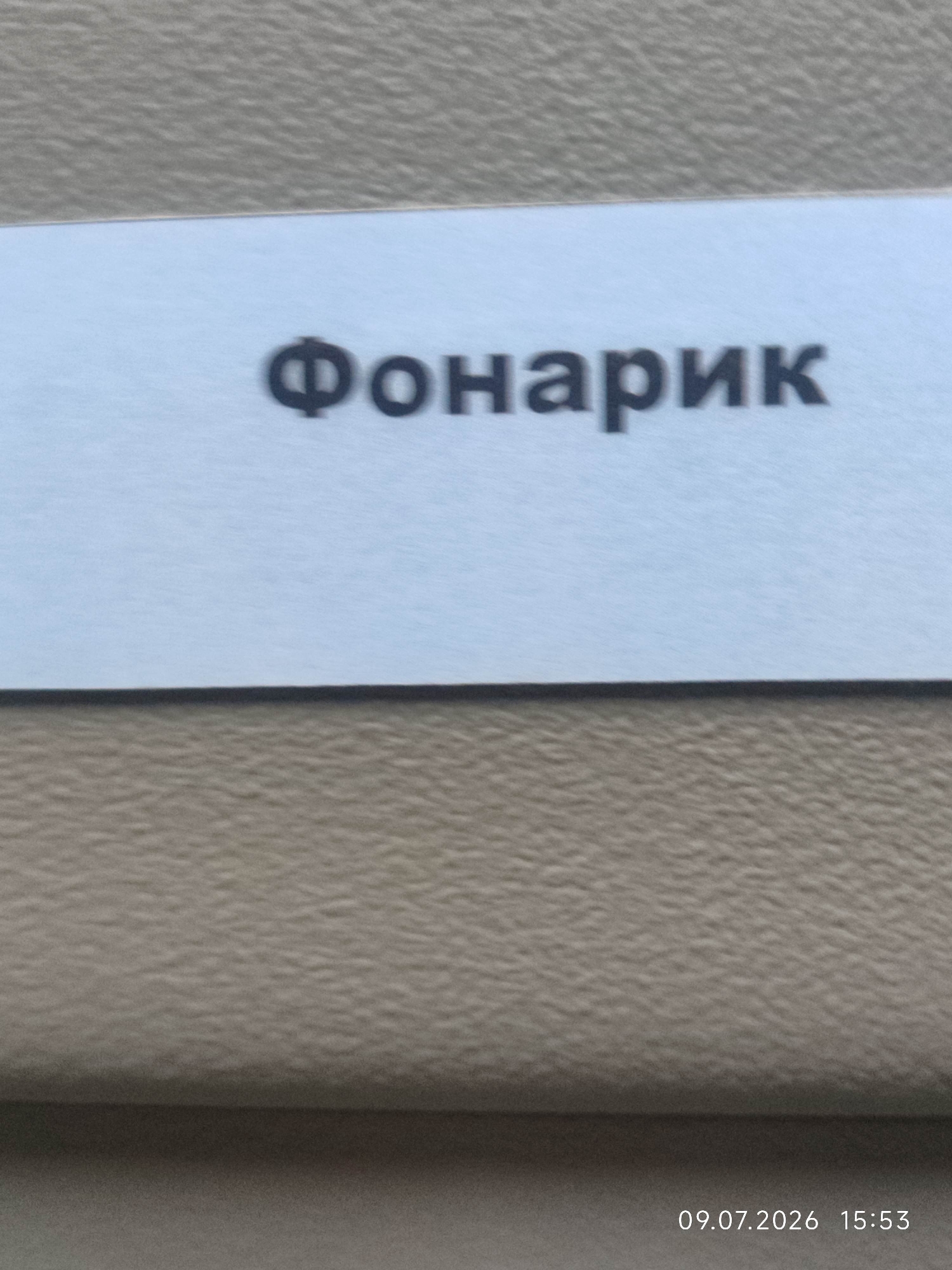 The image displays a light blue rectangular label with the Russian word "Фонарик" (Fonarik), which translates to "flashlight," printed in black, bold, sans-serif font. The label is positioned horizontally against a textured, beige surface, possibly a wall or furniture. The bottom right corner of the image includes a timestamp: "09.07.2026 15:53." There are no people or other discernible objects in the frame. The image is cropped such that only the label and a portion of the background surface are visible.