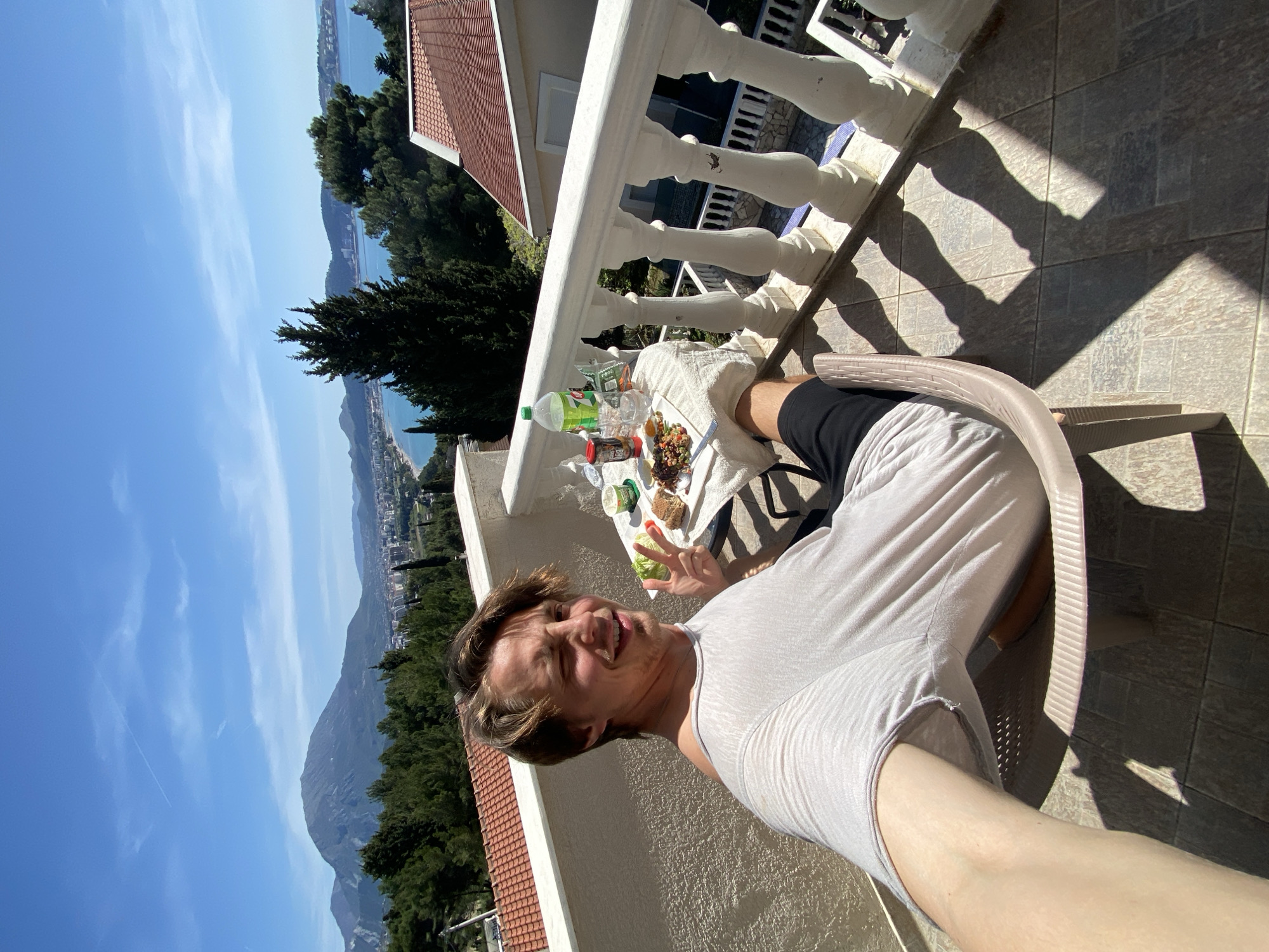 A person is taking a selfie on a balcony in Šušanj, Montenegro. The person, wearing a white shirt and black shorts, is seated in a beige plastic chair. A table covered with a white cloth has food, drinks, and containers on it. Behind the balcony, there is a view of the city, mountains, the sea, and a blue sky. The balcony has a white balustrade. The floor is tiled with gray square tiles. Shadows from the balustrade are cast onto the floor.