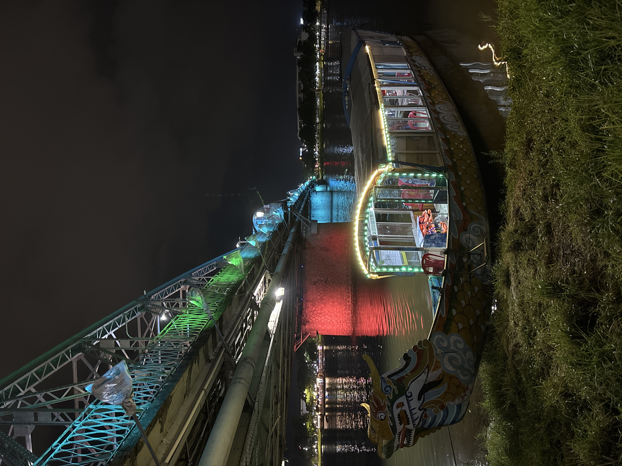 This image depicts a nighttime scene in Phú Xuân Ward, Vietnam, with a focus on a dragon boat moored on a riverbank, illuminated by strings of yellow lights. The boat's ornate dragon head prow is visible, painted in vibrant blues, yellows, and reds. Beyond the boat, a large bridge spans the river, lit with green and red lights. The bridge's structure is a complex lattice of metal girders. In the background, the opposite bank of the river is lined with trees and buildings, also illuminated, with lights reflecting on the water's surface. The sky is dark, with some faint clouds. The foreground consists of a grassy bank. There are no people clearly visible in the image, though the interior of the boat suggests it is set up for passengers.