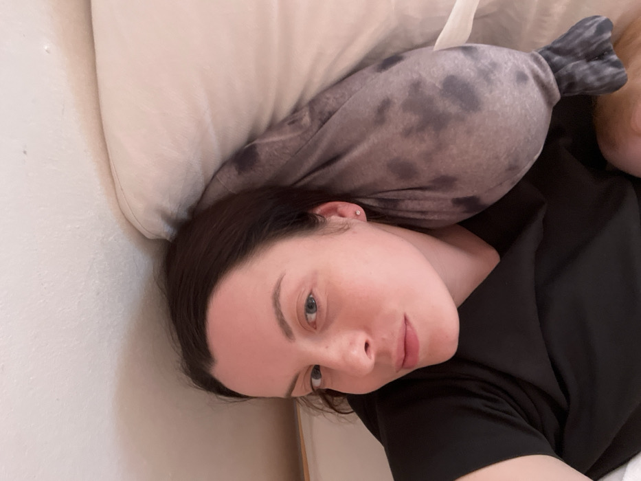 A female subject is depicted lying in an indoor setting, identified as Hohenbrunn, Germany, likely a bedroom. She has dark brown hair, fair skin, and blue eyes, and is wearing a dark, short-sleeved top. A small stud earring is present in her visible left ear. The subject is positioned on her left side, looking directly towards the camera with a neutral expression. To her right, a large gray stuffed animal with irregular dark spots, resembling a seal, rests partially on a light-colored pillow. A plain, light-toned wall is visible in the upper left background.