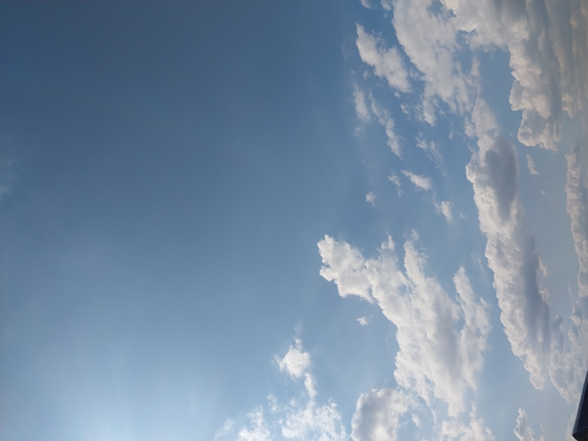 The image shows a clear blue sky with scattered white cumulus clouds. The sun is visible in the upper left corner, casting a slight glare. The bottom of the frame shows a sliver of a dark, sloped structure, possibly a rooftop, suggesting an outdoor scene in Nizhneye Myachkovo, Russia.