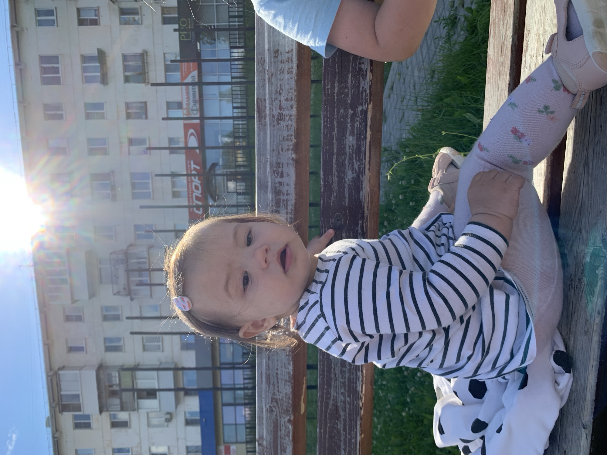 A young child, likely a toddler, sits on a weathered wooden park bench outdoors on a sunny day. The child is wearing a striped long-sleeved shirt, patterned tights, and pink sandals. A blanket with a black and white polka dot pattern is draped over their lap and the bench to their left. In the background, a multi-story apartment building is visible, with balconies and windows. Sunlight flares across the scene. This appears to be a residential area in the city of Chaikovsky, Russia, given the architecture of the building and typical urban park setting. Another person, only their arm and shoulder visible, is seated to the child's right.