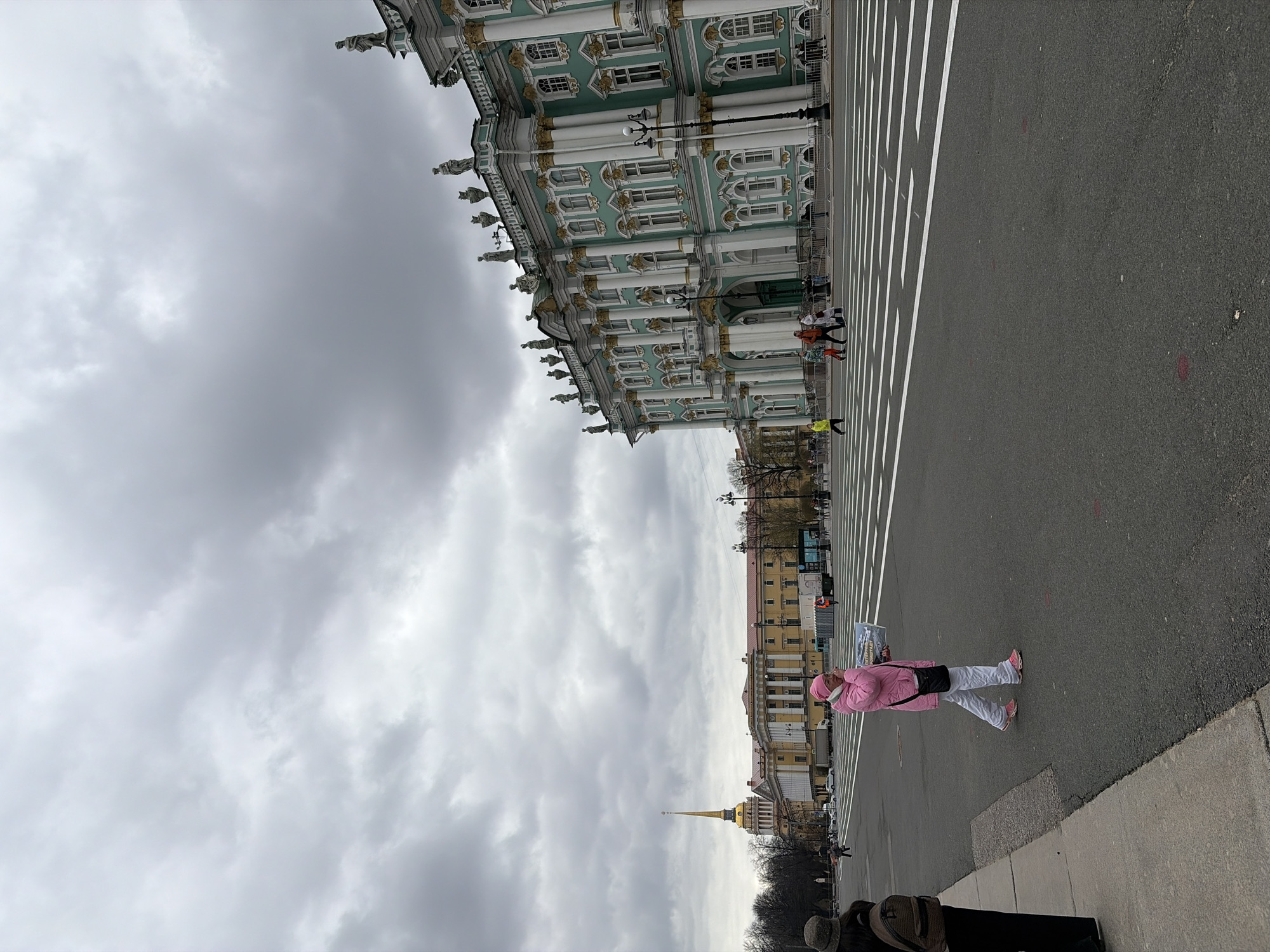 The image captures an expansive urban scene in Saint Petersburg, Russia, under a uniformly overcast, grey sky. The majestic Winter Palace, part of the State Hermitage Museum complex, dominates the left side of the frame. It displays distinctive Baroque architecture with light green walls, classical white columns, and intricate gilded details, including statues adorning the roofline and ornate window frames.
In the mid-ground and foreground, a broad asphalt thoroughfare or square stretches across the scene. This area is marked with a striking pattern of wide white stripes, likely indicating a large pedestrian crossing or decorative street design.
A prominent person is visible in the mid-right foreground, dressed in a vibrant pink puffer jacket, light-colored pants, and pink sneakers, carrying a dark shoulder bag. This individual is holding a sign that clearly reads "FREE WALKING TOUR," followed by a website address "st-petersburg-tours.ru" and an Instagram handle "@st_petersburg_tours." Several other smaller figures are scattered in the distance, near the palace and further along the road, one of whom is wearing a bright yellow jacket.
In the background, on the right, additional city buildings are visible, most notably featuring a golden spire characteristic of famous Saint Petersburg landmarks, likely the Admiralty Building. Leafless trees suggest the season is late autumn, winter, or early spring. The heavy cloud cover results in muted, dull daylight across the entire scene. The activity appears to be an individual advertising or conducting a walking tour in a bustling historic city square.