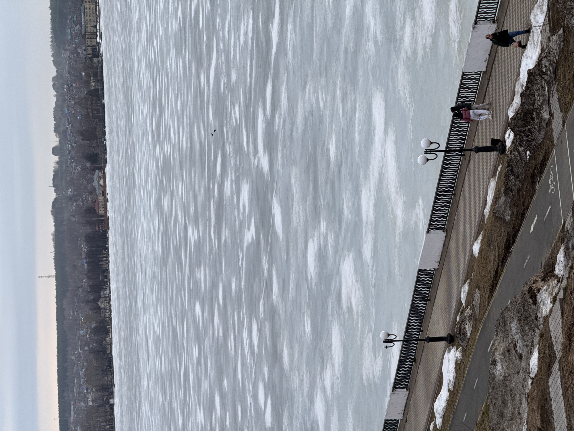 The image depicts a frozen body of water, possibly the Votkinsk Reservoir, adjacent to a paved embankment in Votkinsk, Russia. The water surface exhibits a pattern of alternating light and dark streaks, indicative of partially melted or unevenly frozen ice. Along the embankment, there are pedestrian walkways and a dedicated bicycle path. Several people are visible on the embankment. One person is walking alone, while a couple is seen standing together. Street lamps are positioned along the embankment. In the background, there is a line of trees and some buildings. The sky is overcast. Patches of snow are observed on the embankment and grassy areas.